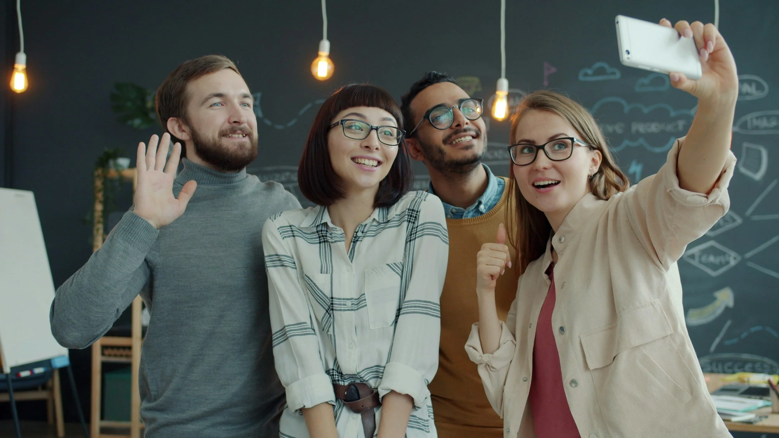 Four people taking a group selfie in an office setting, smiling and making gestures, with a chalkboard wall and hanging light bulbs in the background.