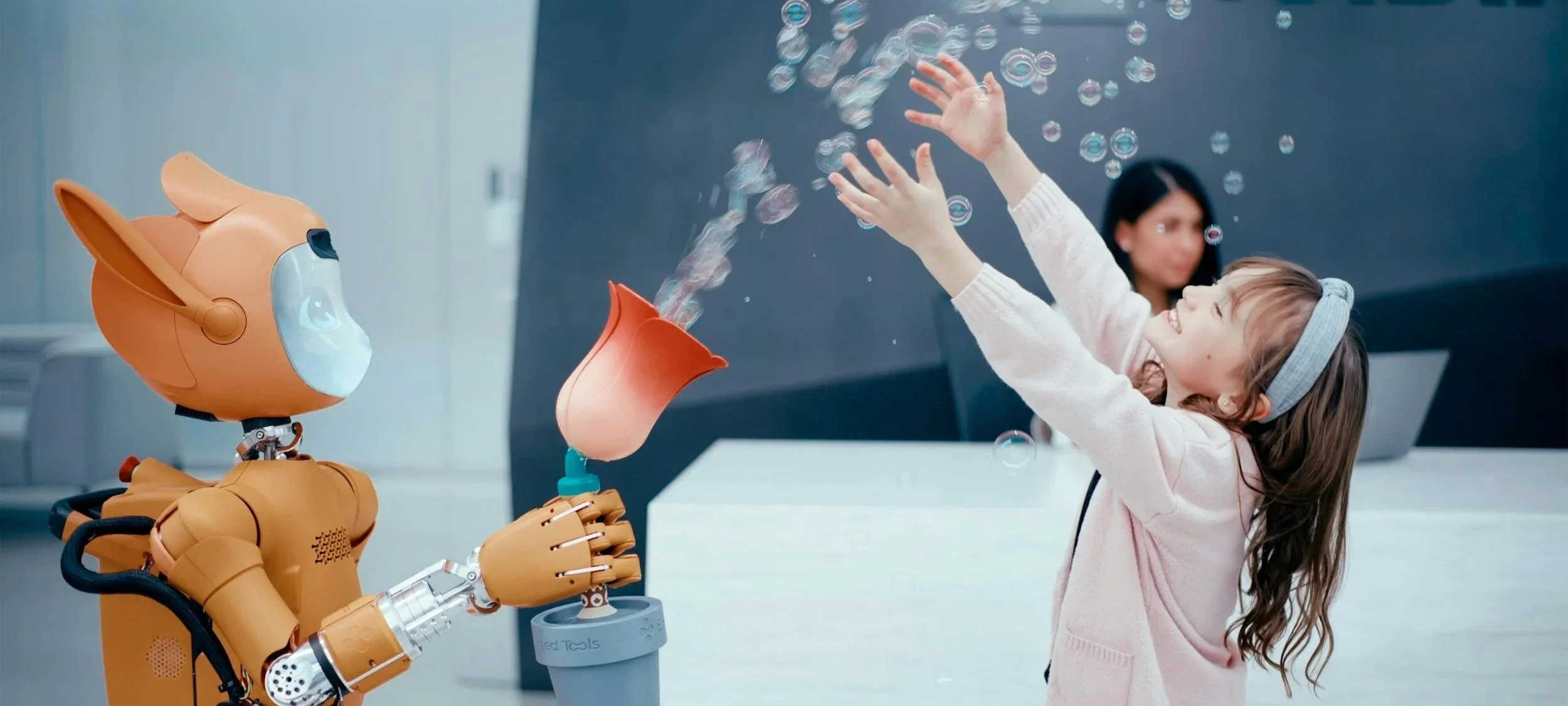 A young girl, smiling and happy, with a headband reaching out to a robot with a dog's head, who is holding a cup with bubbles emerging from it.