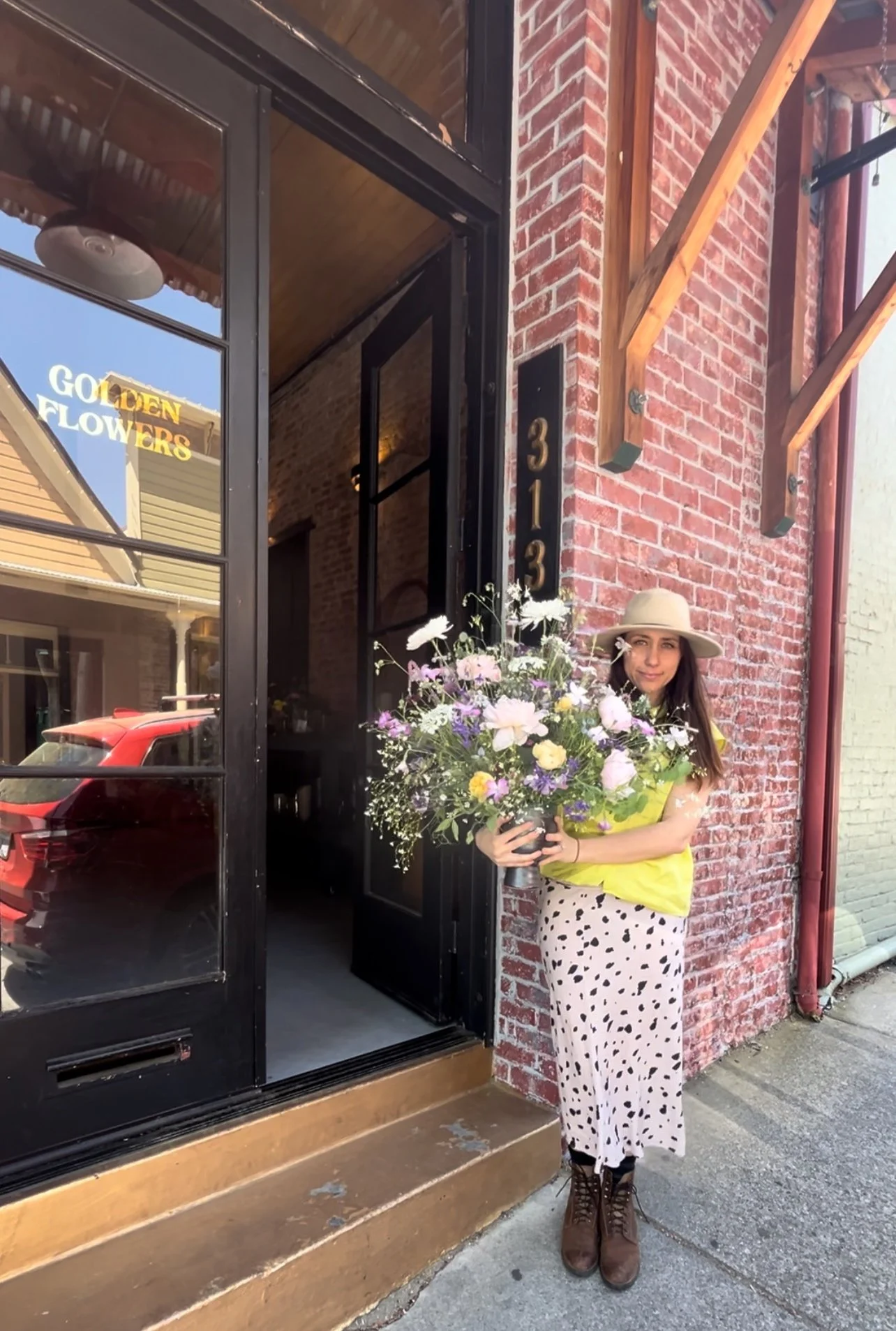 A woman standing outside a brick building holding a large bouquet of flowers, wearing a beige hat, yellow top, white skirt with black polka dots, and brown boots.