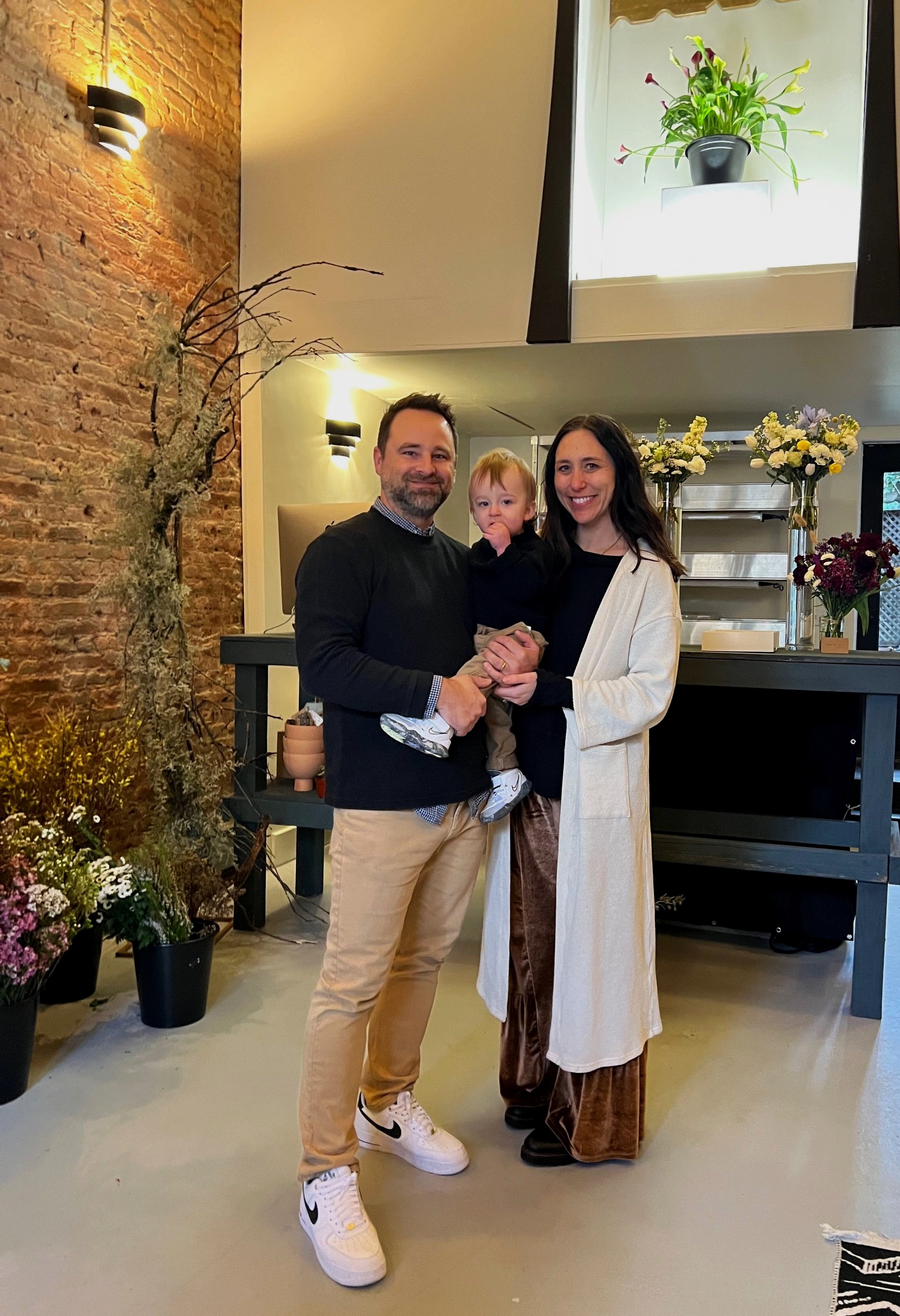 A family of three stands together in a warmly decorated indoor space, smiling at the camera. The man has a beard and wears a black sweater, tan pants, and white sneakers. The woman has long dark hair and wears a white coat over dark clothing. The child has red hair, wears a black top, and is held by the man. The background features potted plants, flowers, and a brick wall.