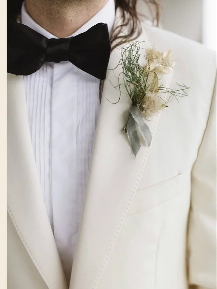 Close-up of a groom wearing a white tuxedo with a black bow tie and a boutonniere consisting of dried flowers and greenery on his left lapel.