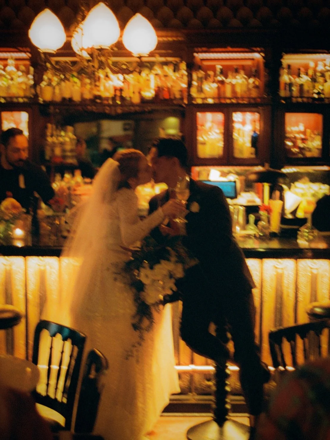 A wedding couple shares a kiss at a bar in a warmly lit venue, with bar shelves filled with bottles in the background.