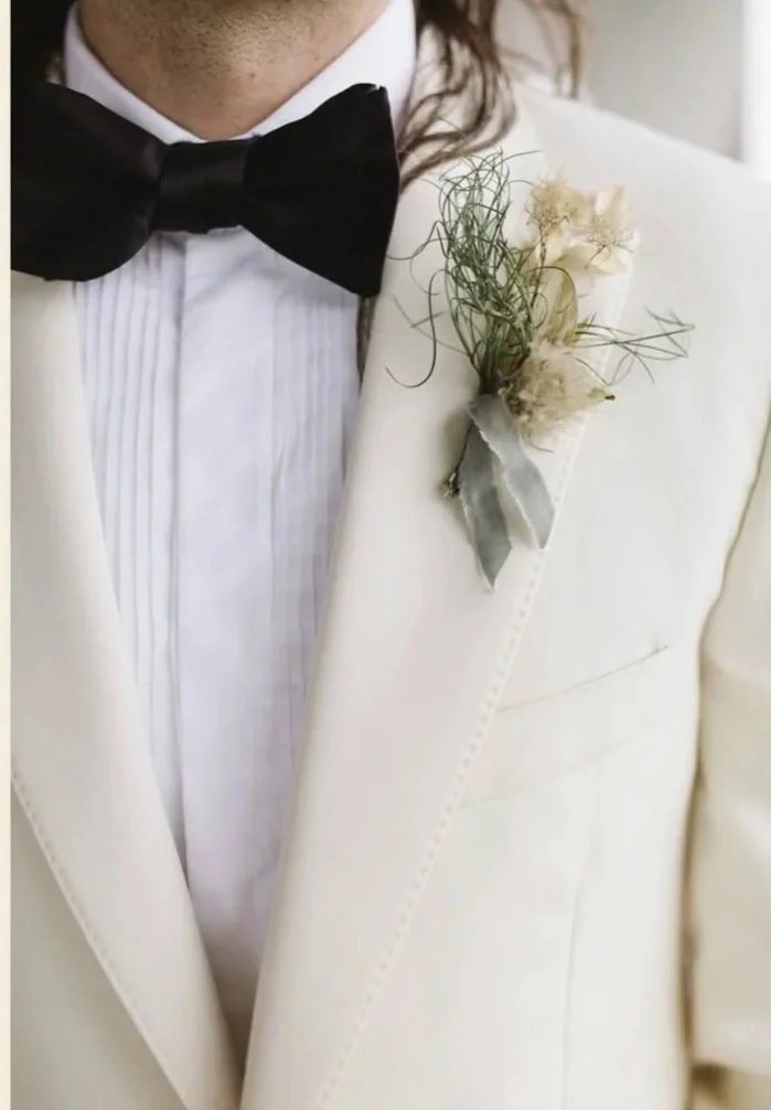 Close-up of a man wearing a wedding flowers in a white tuxedo jacket with a black bow tie and a boutonniere with white flowers and greenery on the lapel.