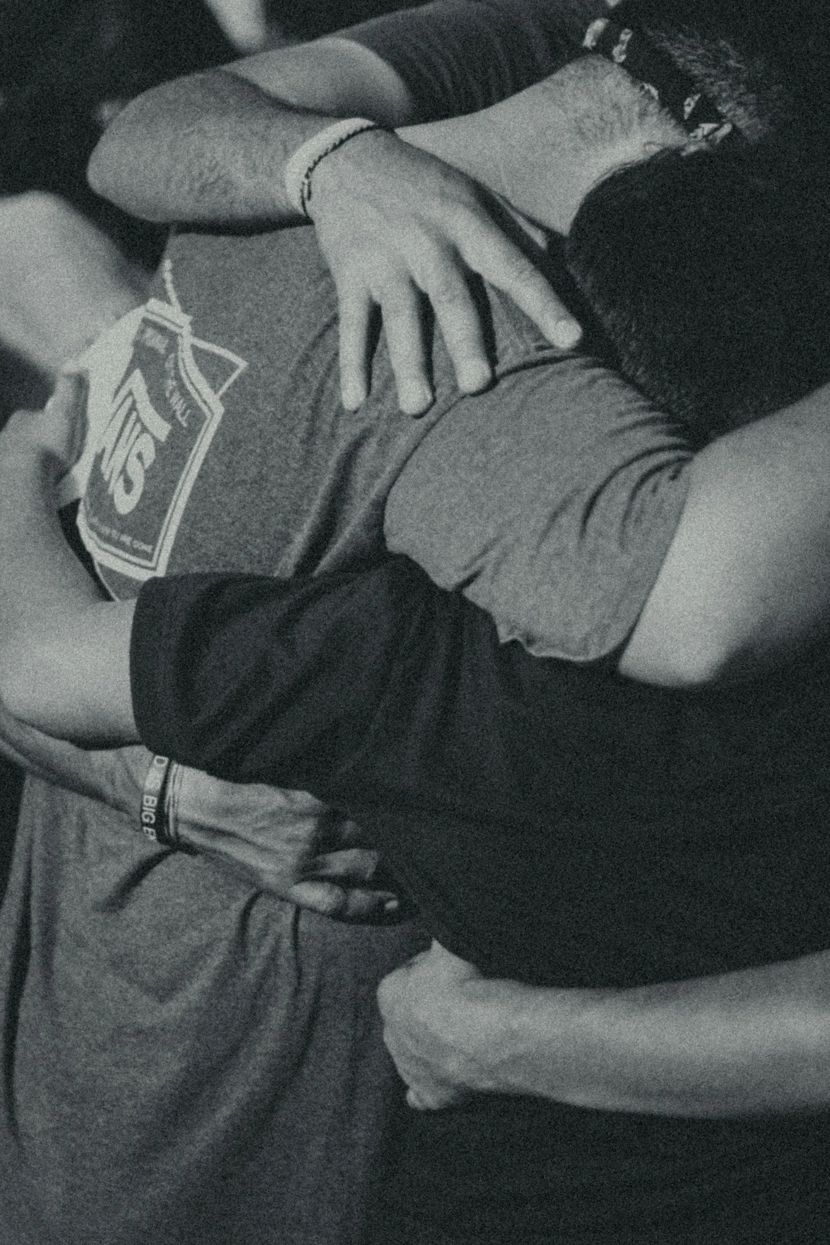 A close-up, black-and-white photo of two people lying on the ground, with one person's hand resting on the other's chest. One person is wearing a shirt with a visible logo and a wristband, while the other is dressed in dark clothing with a patterned sleeve.