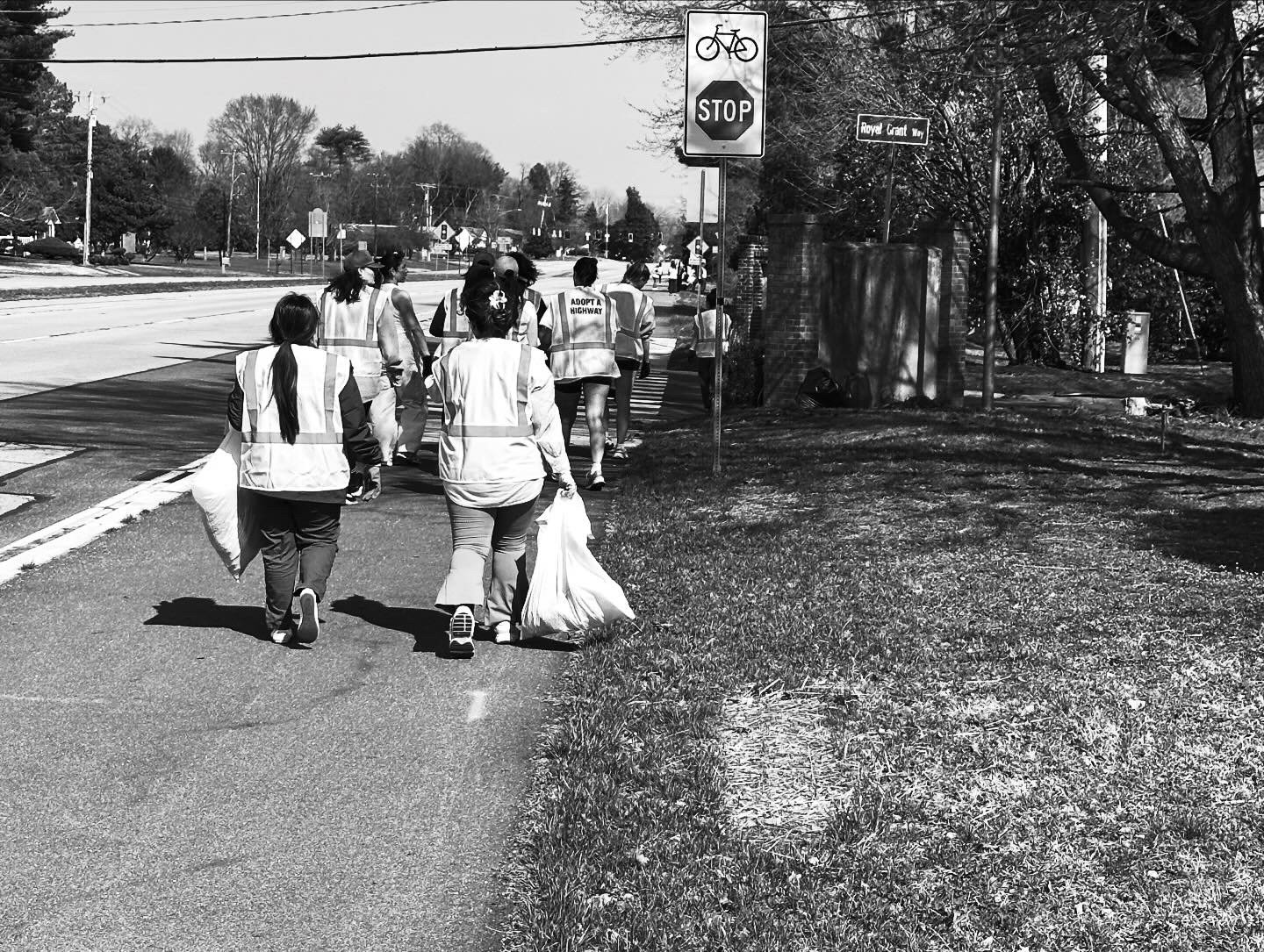 Group of volunteers in reflective vests walking on sidewalk, some carrying bags, near a stop sign, crossing a street in a neighborhood.