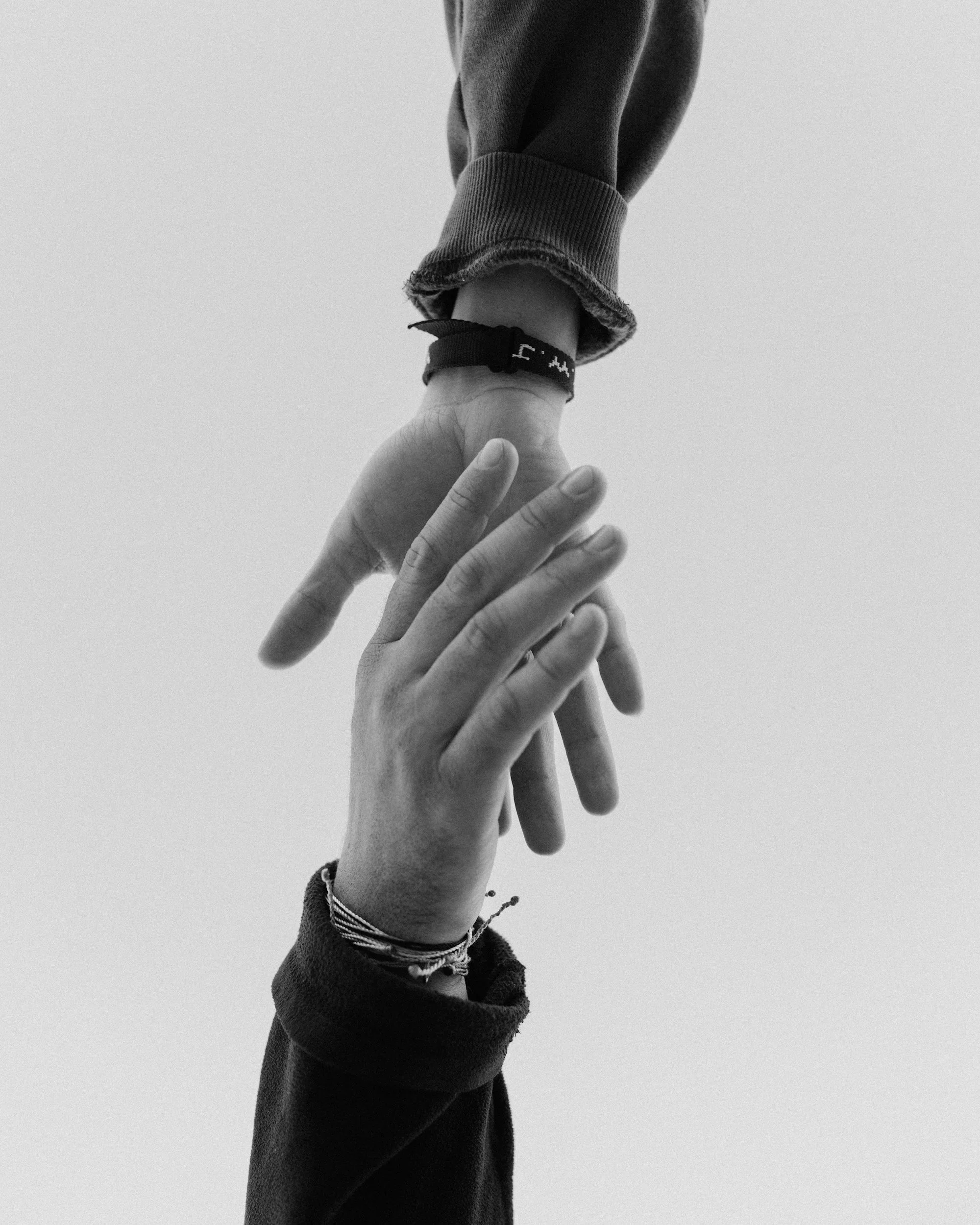 A person's hand reaching out and holding the wrist of another person, with the other person's arm extended vertically. The individual being held wears a dark long sleeve. The image is in black and white.