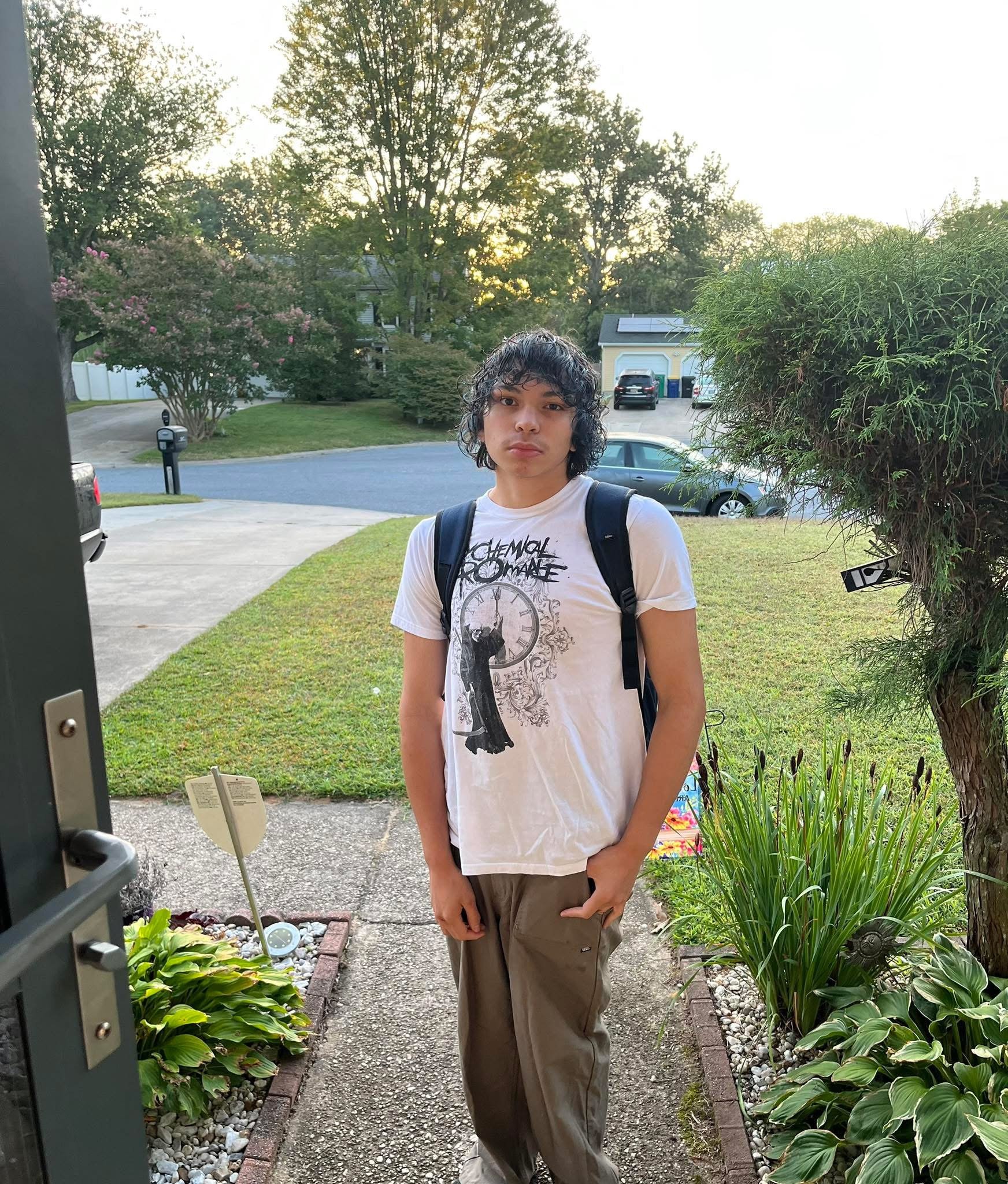 A young man with curly hair and light skin standing outside in a residential area during sunset. He is wearing a white graphic t-shirt, khaki pants, and a backpack. There are trees, bushes, a driveway with parked cars, and houses visible in the background.