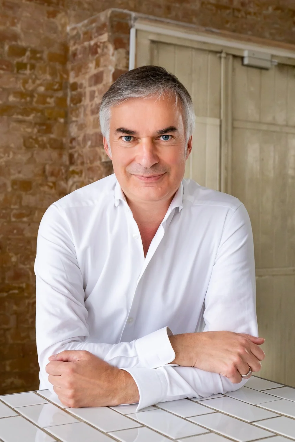 Peter John Mahrenholz in a white shirt sitting at a tiled counter, with a brick wall and a wooden door in the background.