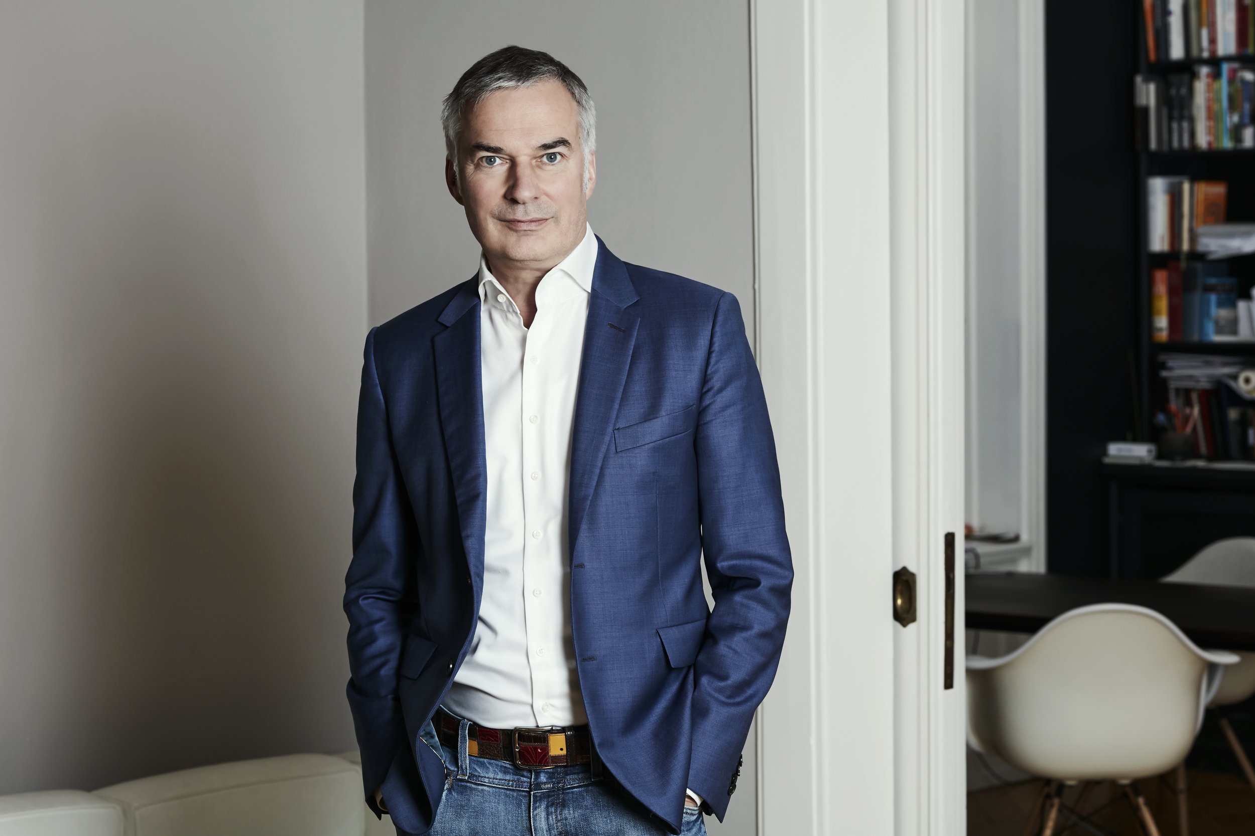 Peter John Mahrenholz in a blue blazer and white shirt standing with his hands in his pockets in a well-lit room with a bookshelf and a white chair in the background.