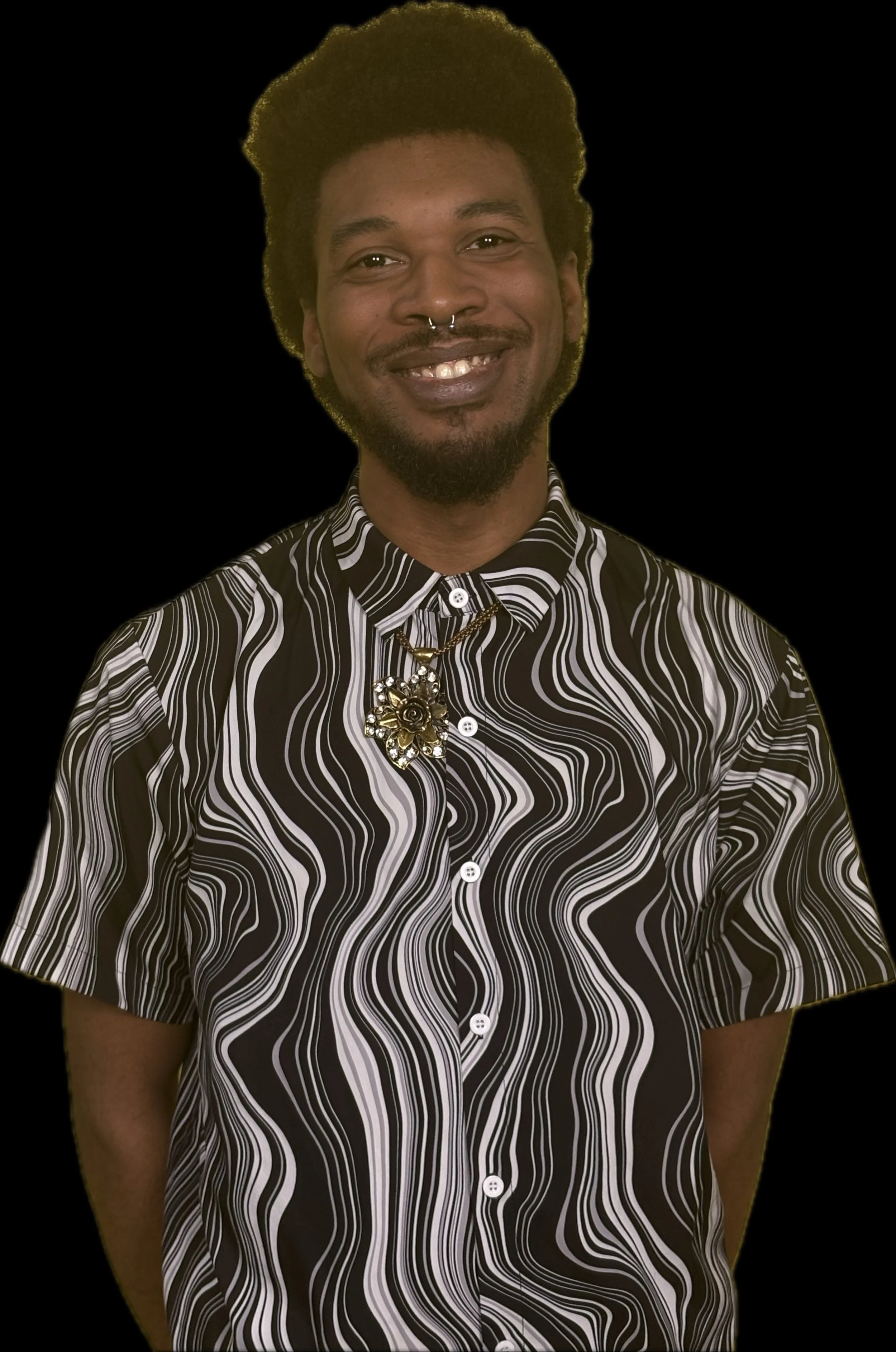 A smiling man with a beard and a septum piercing, wearing a black and white swirling patterned short-sleeve shirt and a floral brooch necklace, posing against a black background.
