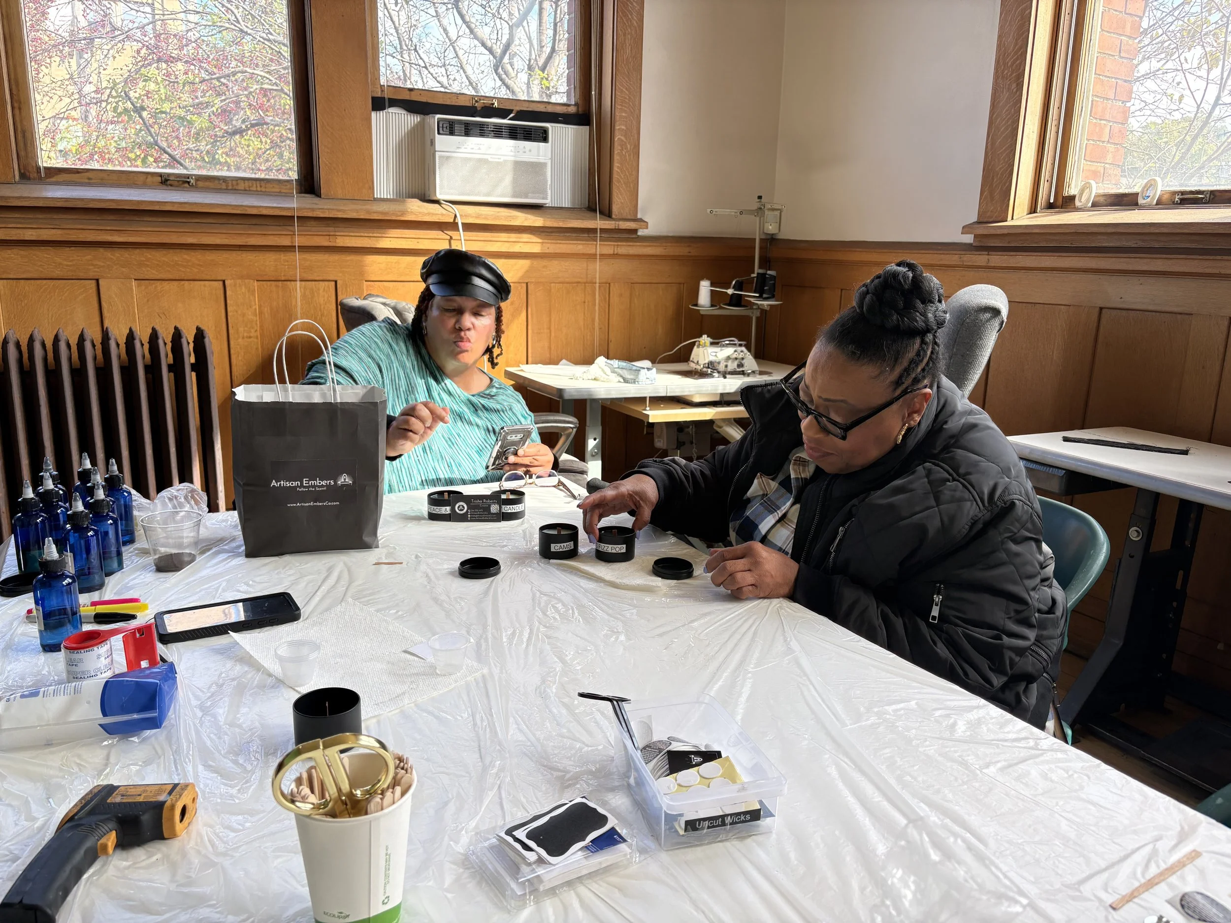 Two women seated at a table crafting with small black containers, surrounded by craft supplies like glue bottles, scissors, and a glue gun, in a room with wooden walls and large windows.