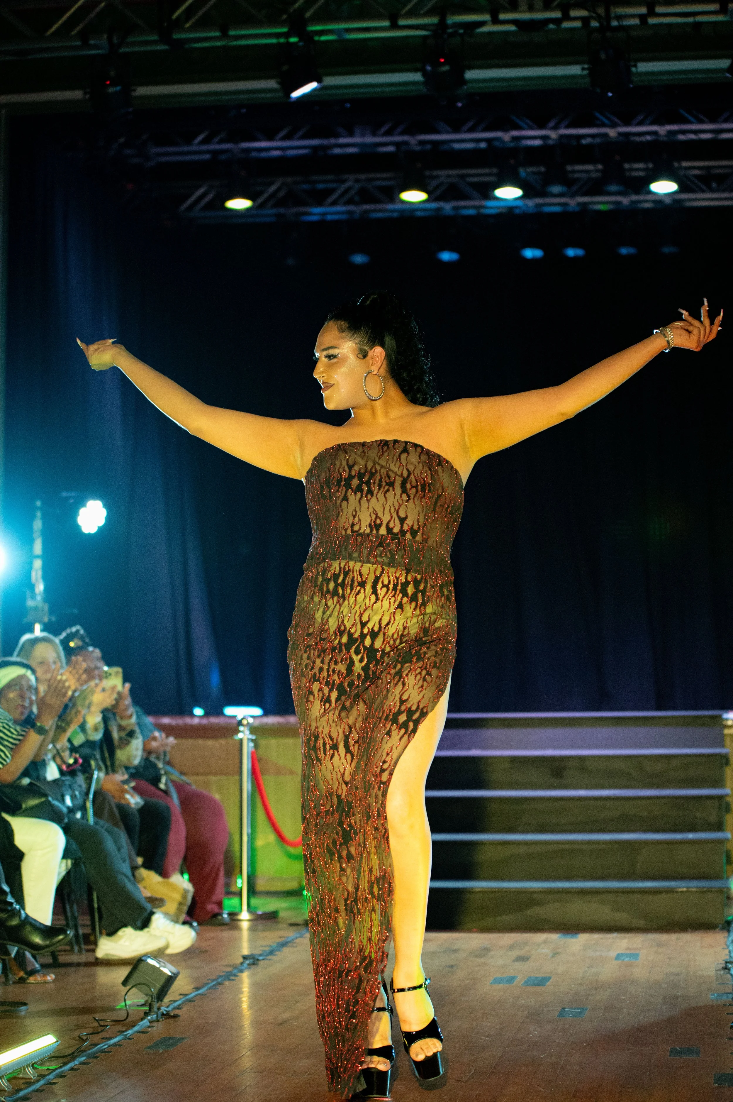 A woman walking down a runway in a strapless, high-slit dress with a fiery pattern, surrounded by an audience taking photos.