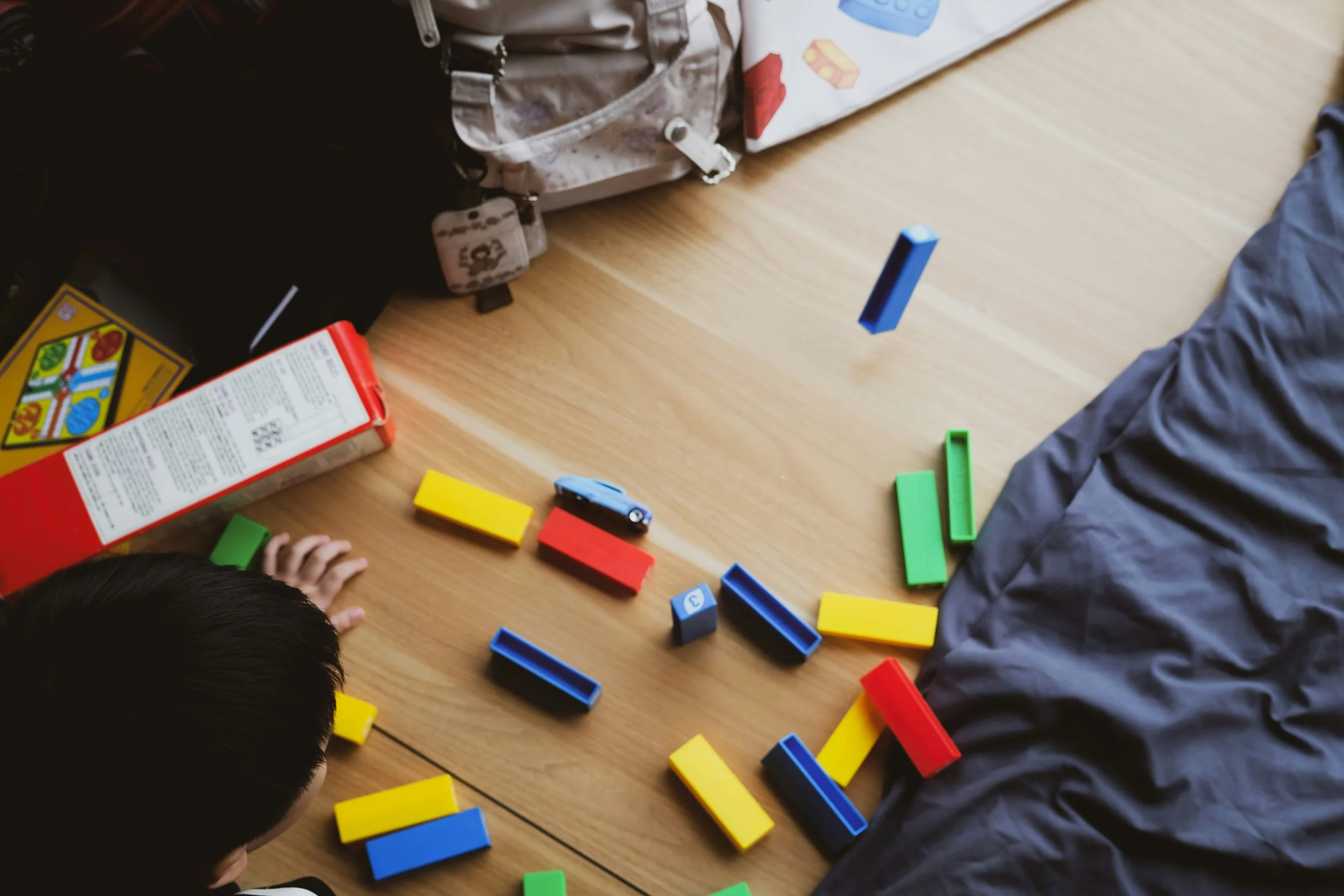 Child playing with colorful rectangular building blocks on a wooden table near a bed and backpack.