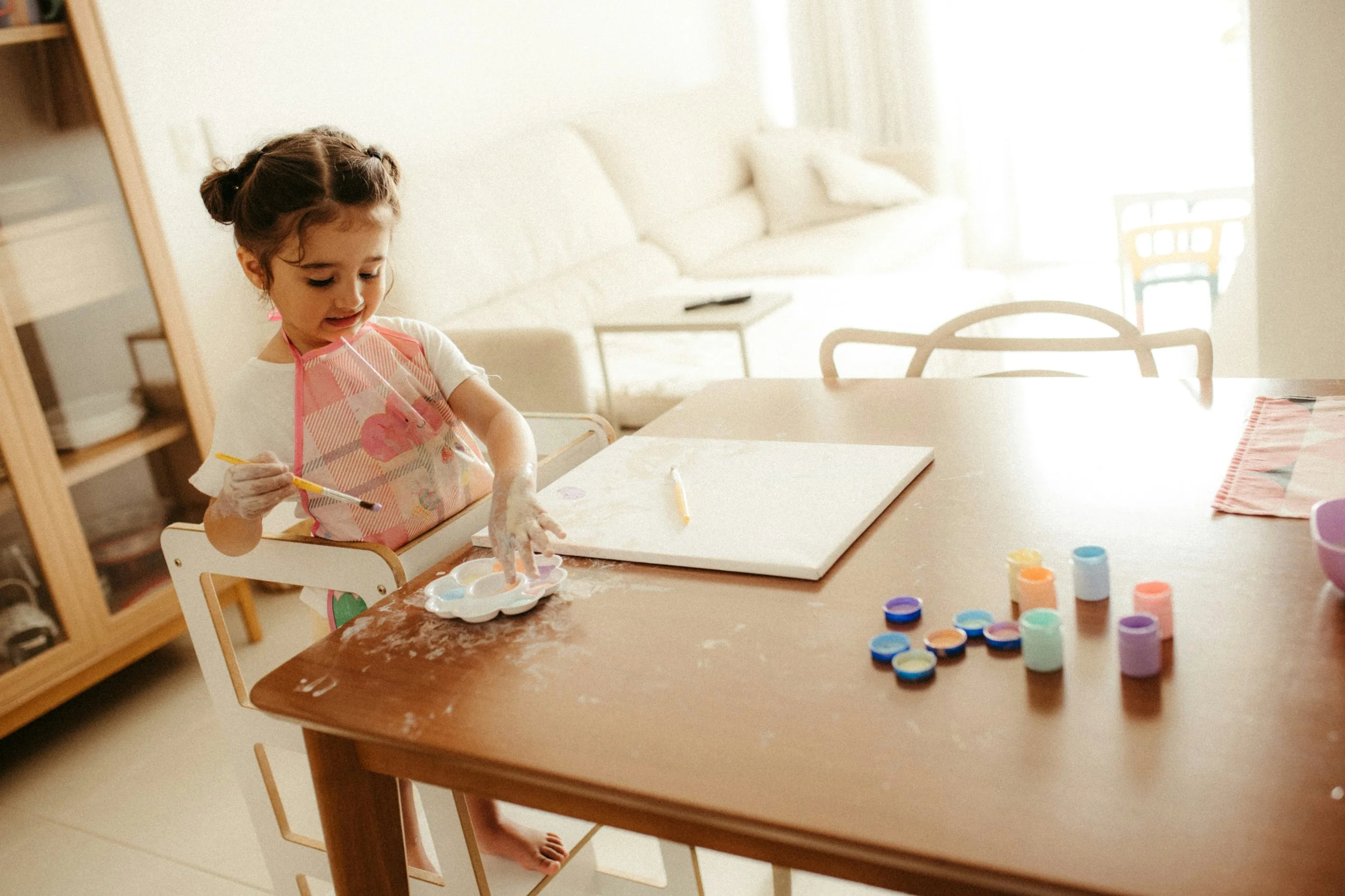 Young girl sitting at a dining table, painting with watercolors, surrounded by paint containers, in a bright living room.