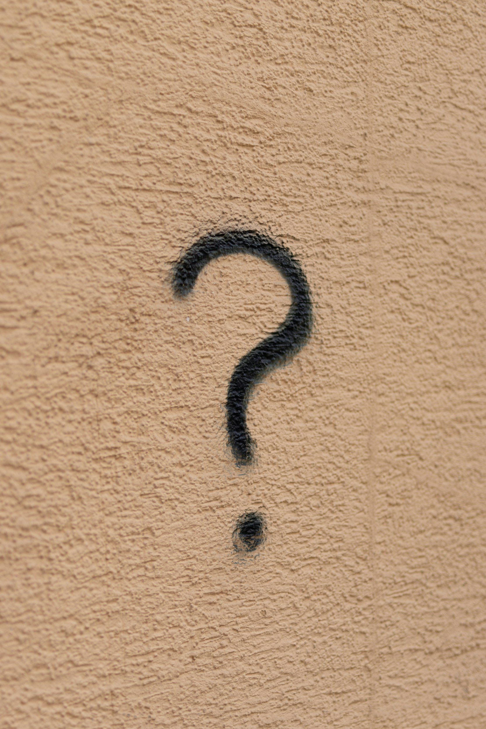 Close-up of a beige textured wall with a black question mark spray-painted on it.