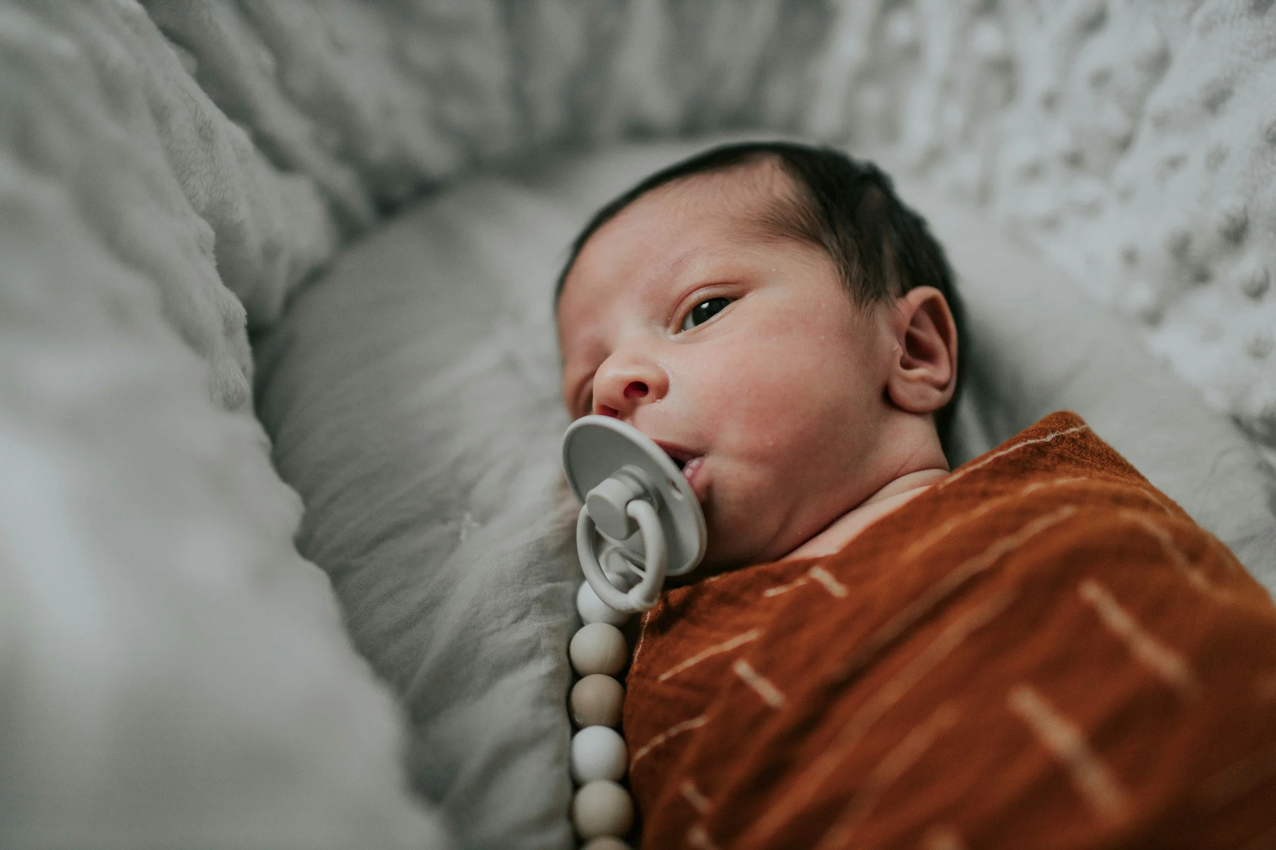 A baby lying on a bed with a pacifier in their mouth, dressed in a brown and orange striped outfit. The baby has short black hair and is looking to the side. The bedding appears soft and gray.