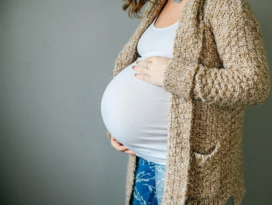 Pregnant woman wearing a white top and a beige sweater, cradling her belly with one hand.