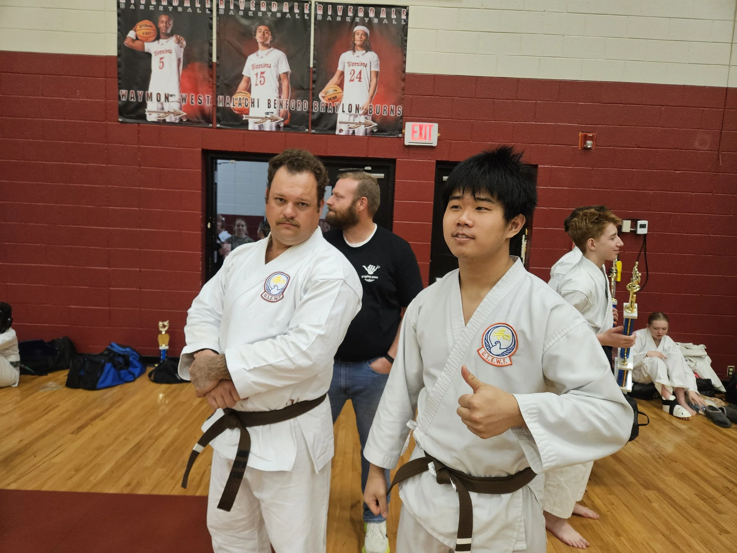 Young martial artists in gi uniforms at a dojo, with trophies in the background and posters of basketball players on the wall.