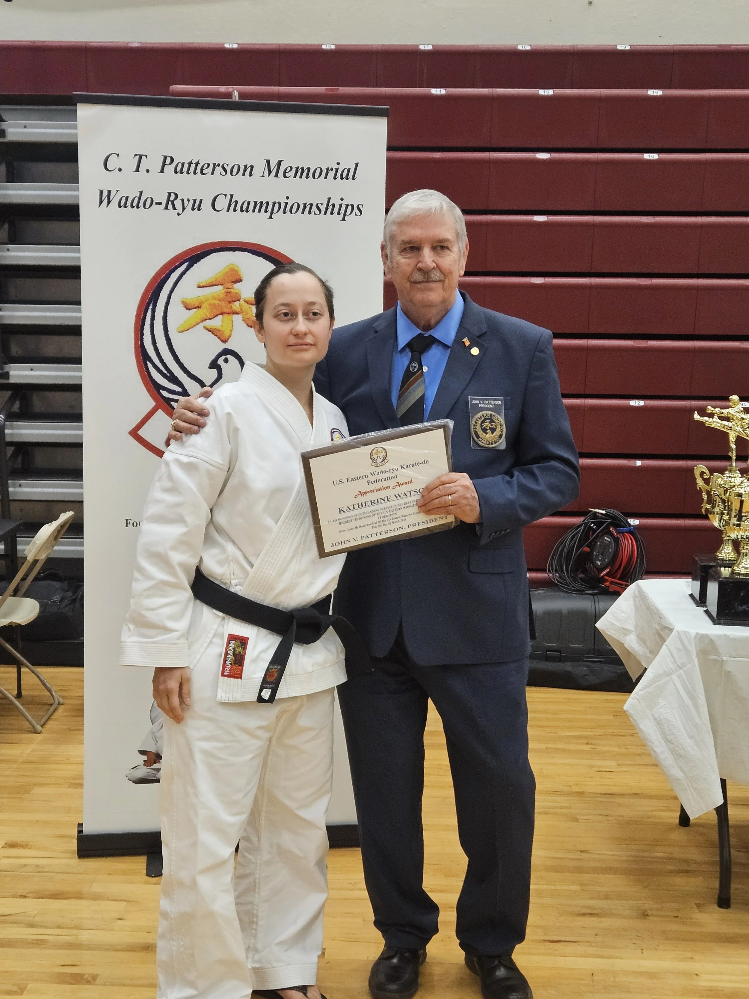 A young woman in a karate uniform holding a certificate stands next to an older man in a suit with a badge. They are in a gymnasium with maroon bleachers and a table with trophies. A banner behind them reads "C. T. Patterson Memorial Wado-Ryu Champio
