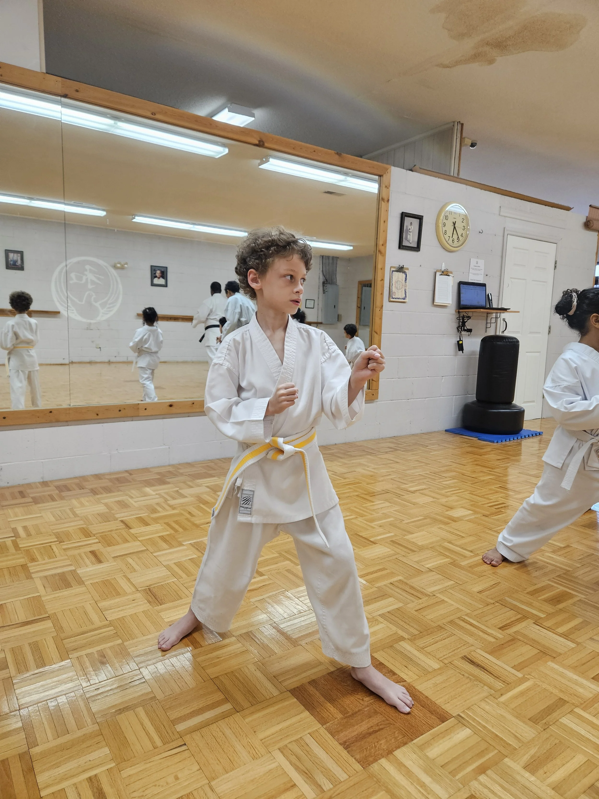 A young boy practicing karate in a dojo, wearing a white gi with a yellow belt, standing in a fighting stance barefoot on a wooden floor, and focusing forward in front of a large mirror.
