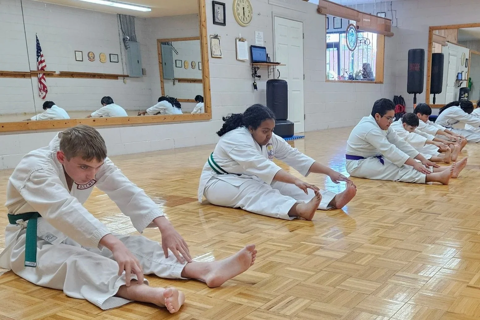 Children in martial arts uniforms stretching on a wooden floor in a dojo, with mirrors and martial arts equipment visible in the background.