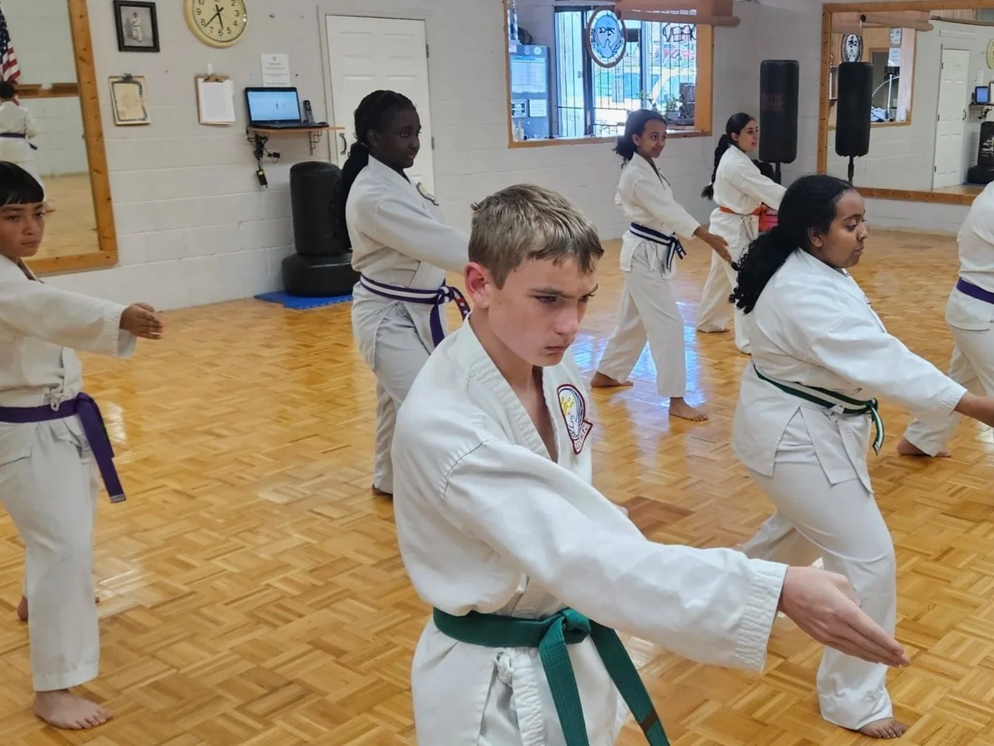 Children practicing martial arts in a dojo, wearing traditional uniforms and colored belts, with a wooden floor and mirrors on the walls.