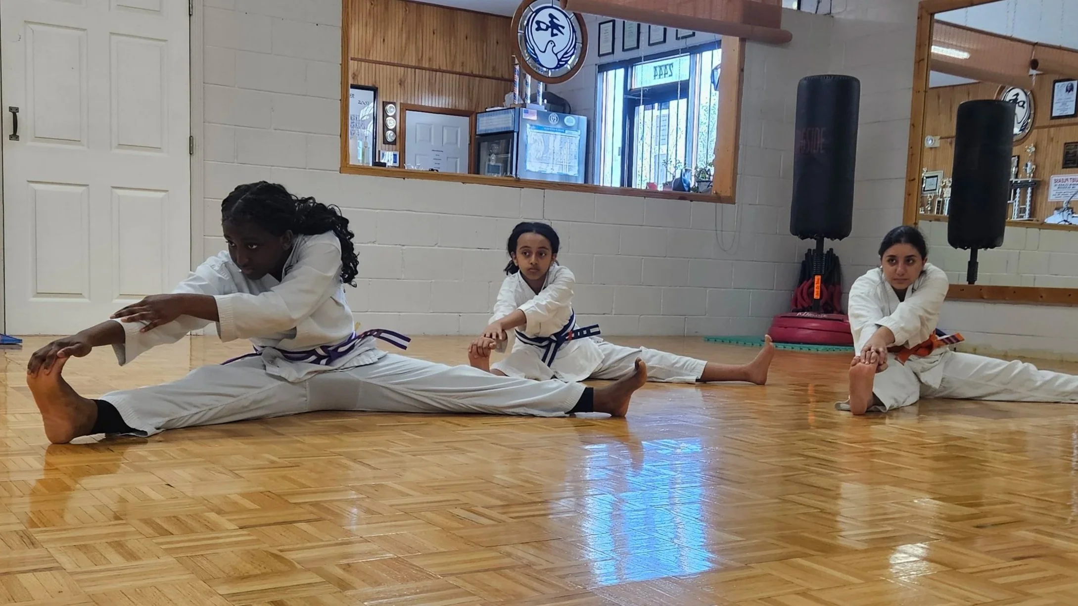 Three young girls in white karate uniforms stretching on a wooden floor in a dojo. They are sitting with legs extended and reaching toward their toes, practicing flexibility.
