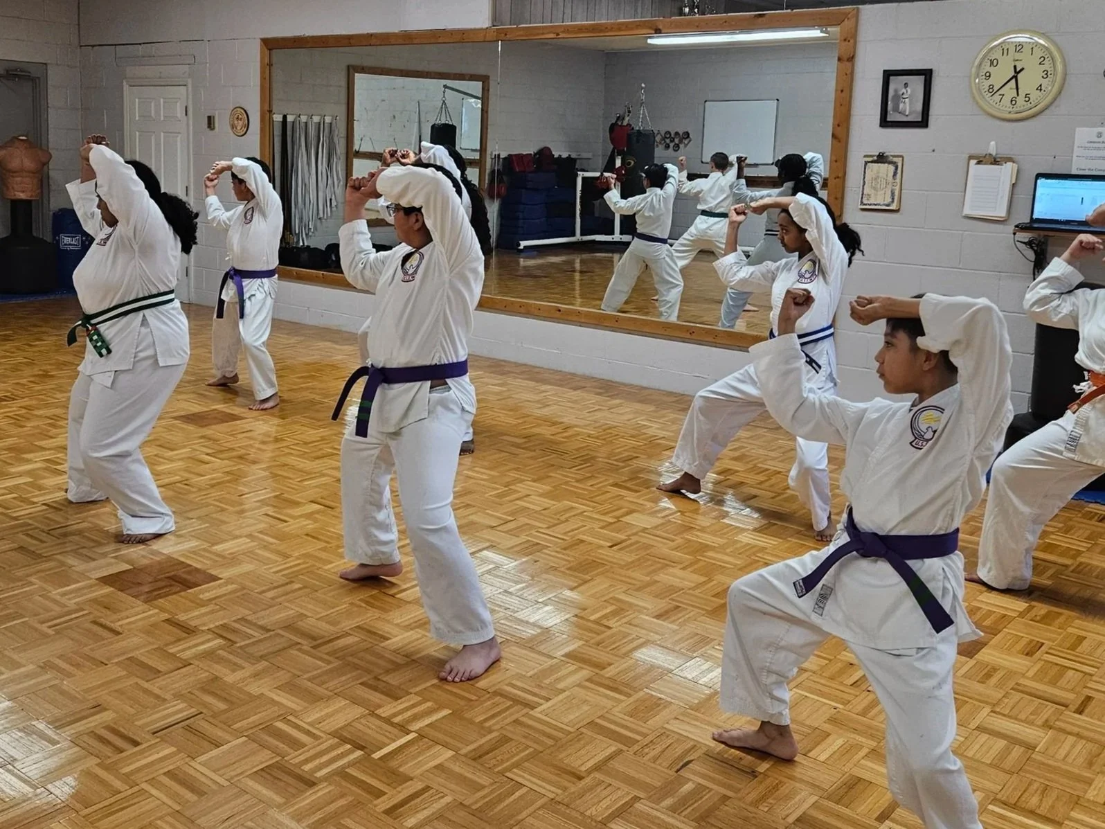 A group of children practicing martial arts in a dojo, wearing traditional uniforms and colored belts, performing a move in front of a mirror.