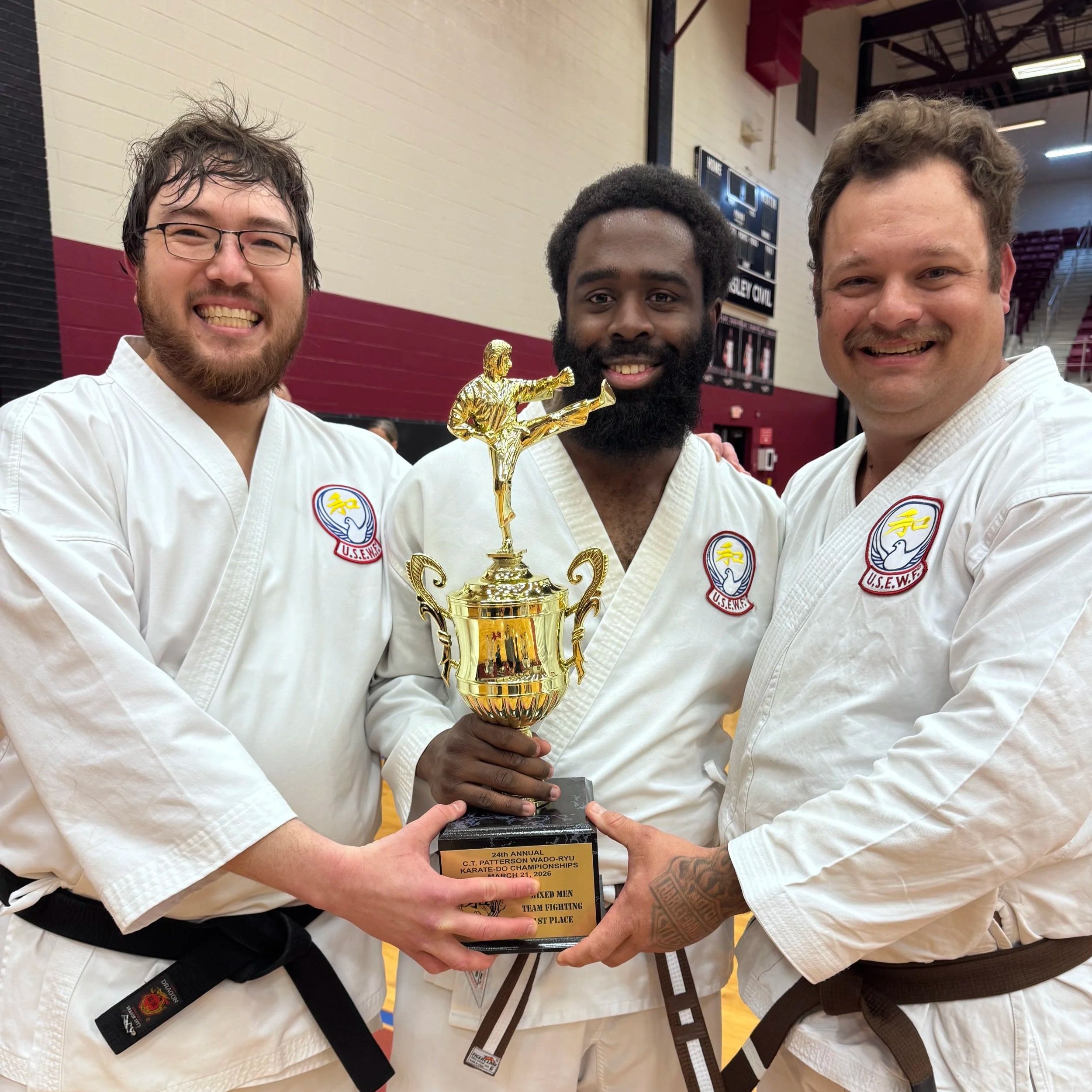 Three men in karate uniforms holding a trophy together in a gymnasium.
