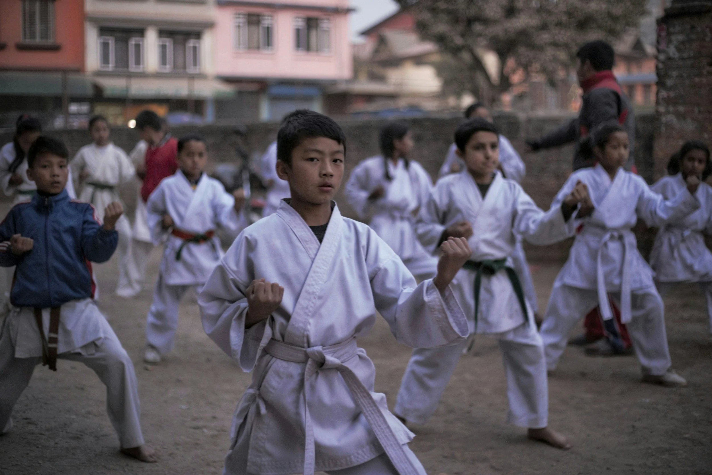A group of children practicing martial arts outdoors, wearing traditional white uniforms (gi) with belts of various colors, in a training session or class.