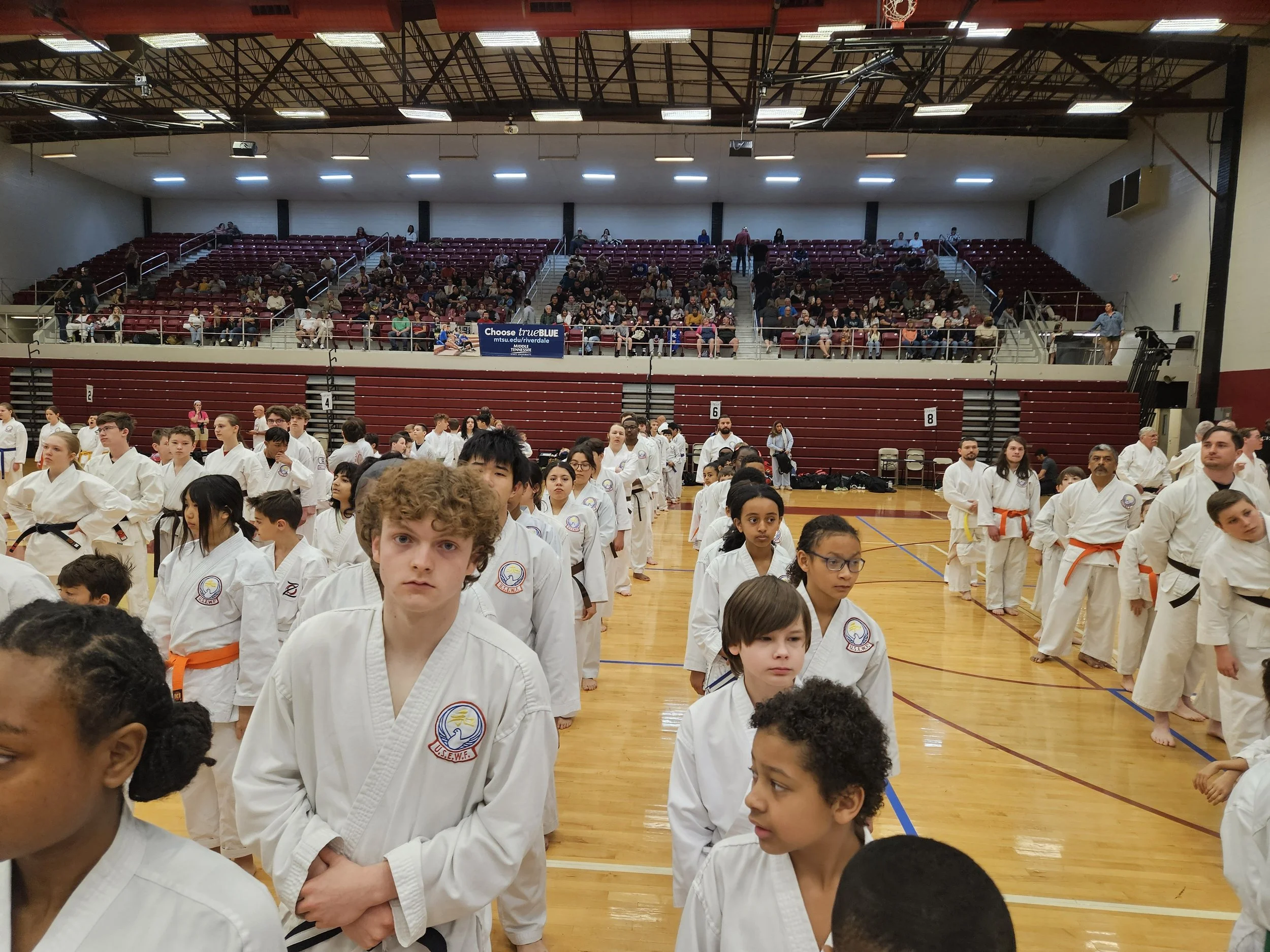 A large group of young martial artists dressed in white uniforms with various colored belts standing in orderly lines on a gymnasium floor for a martial arts event. Spectators seated in the bleachers watch from above.