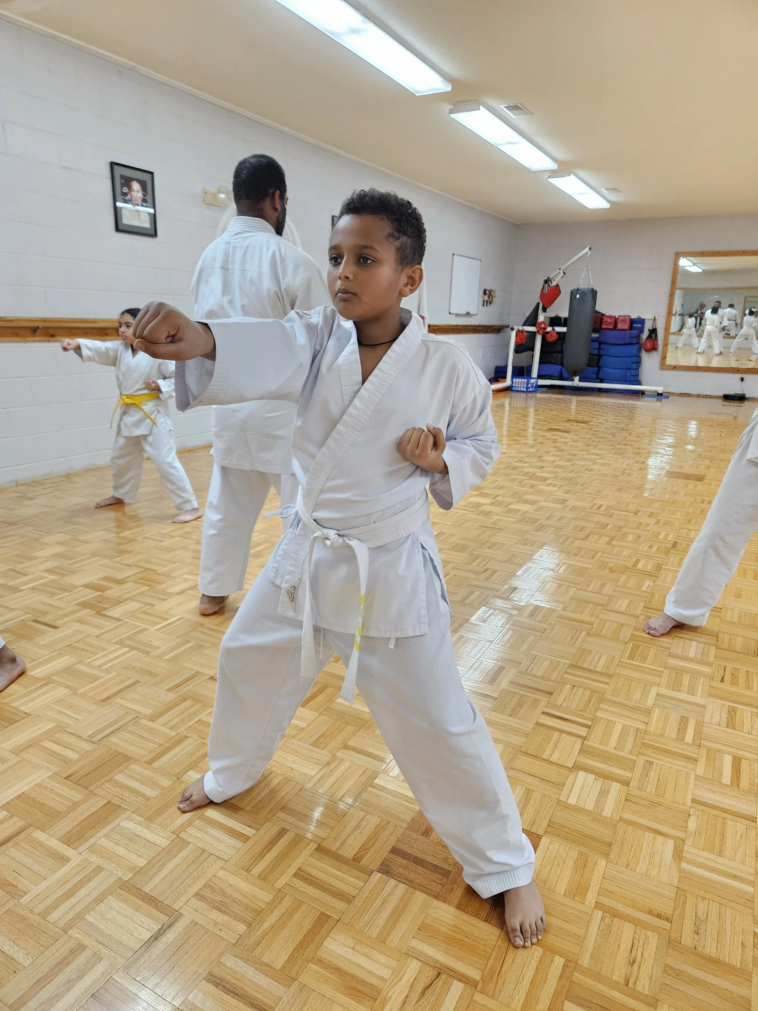 A young boy in a martial arts uniform practicing a punch in a dojo, with others and an instructor in the background.