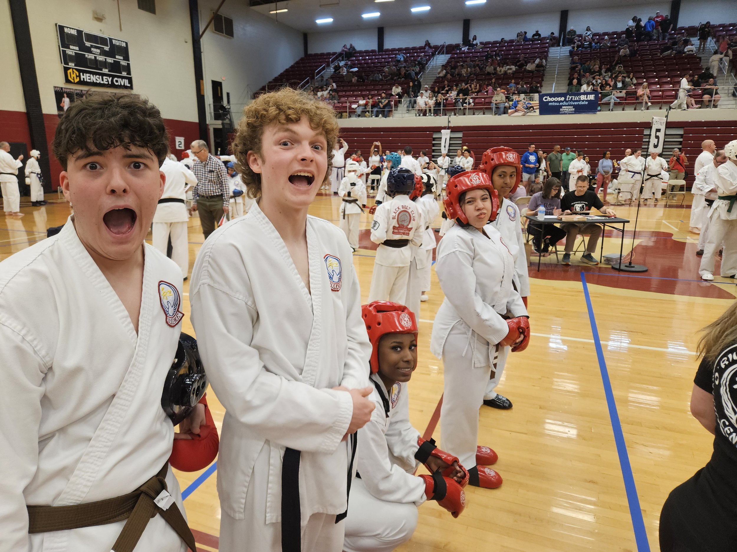 Group of martial artists in uniform at a martial arts tournament, with some wearing red helmets and gloves, standing in a gymnasium with a crowd in the background.