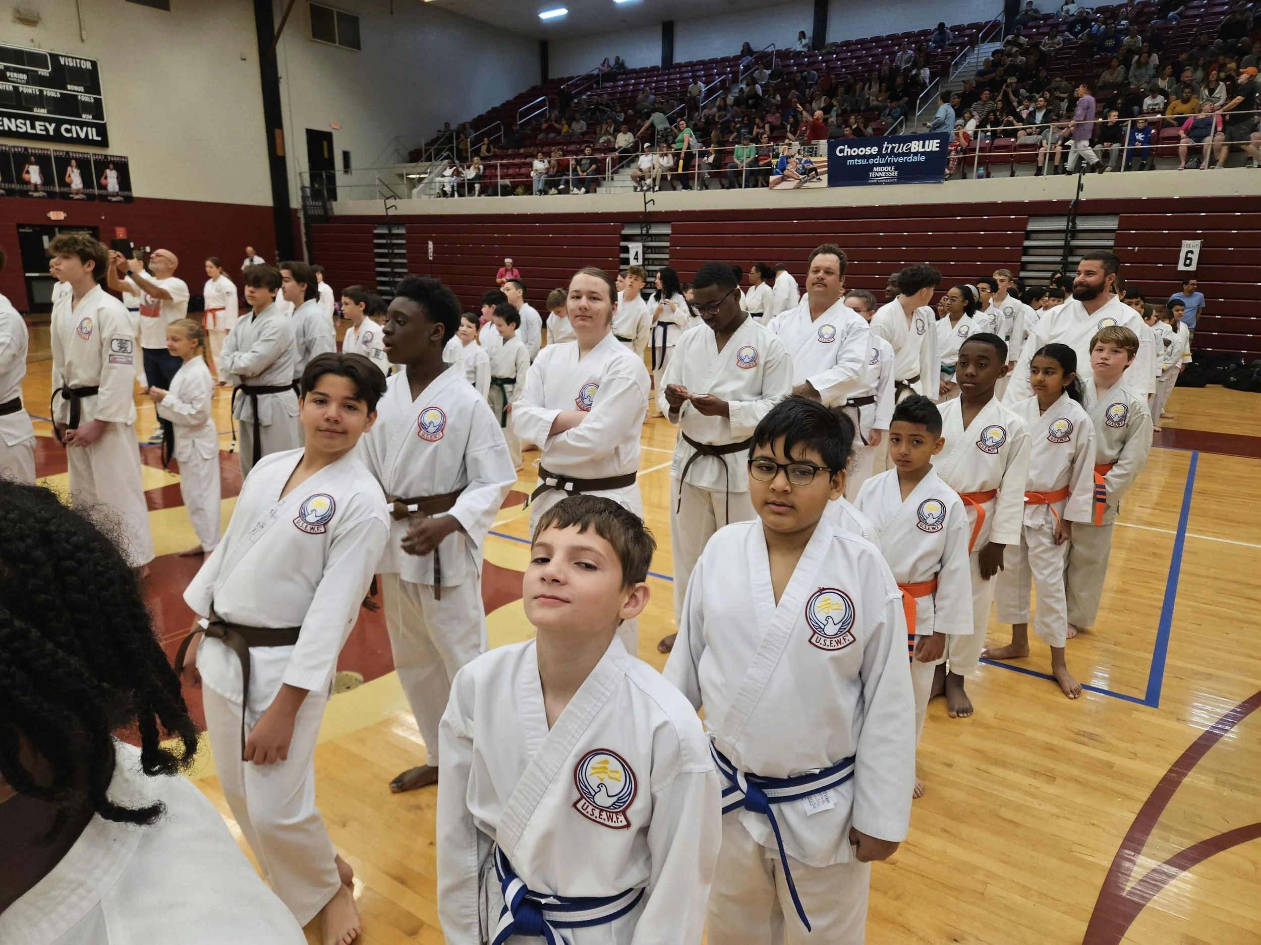 Children and adults in martial arts uniforms standing in line on a gymnasium floor during a martial arts event.