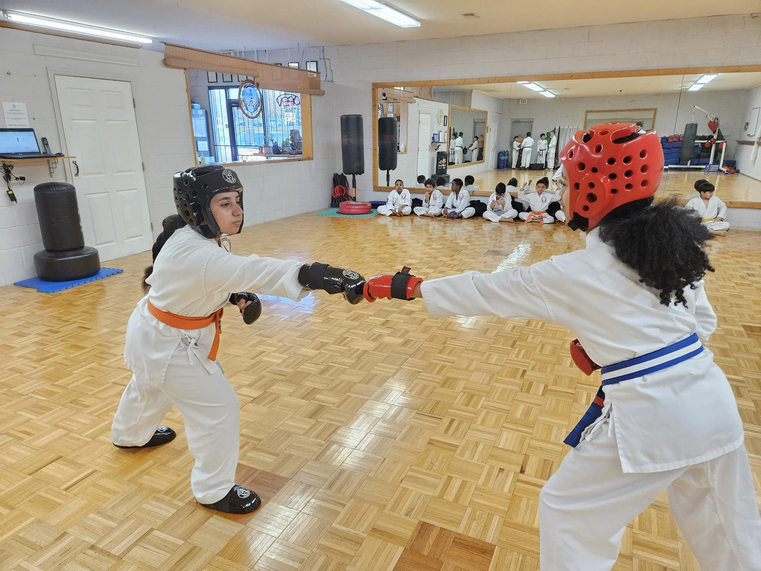 Two young martial artists wearing protective gear, helmets, and white uniforms practicing sparring in a dojo with a wooden floor. Other students sit against the wall in the background.