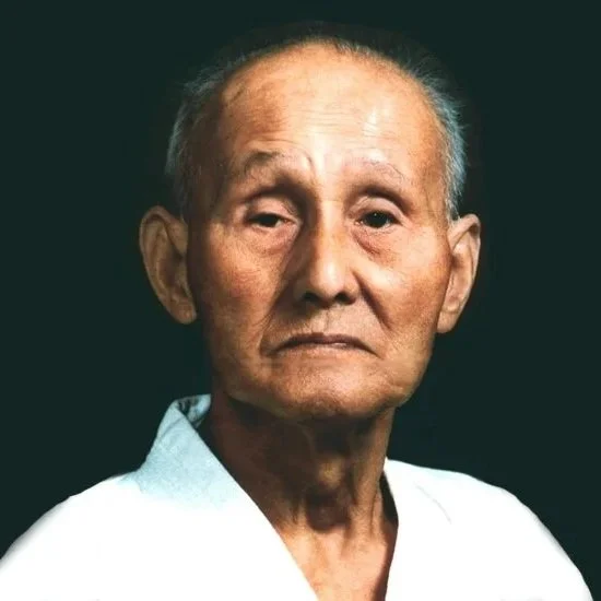Close-up portrait of an elderly man with short gray hair, wearing a light-colored shirt, against a dark background.