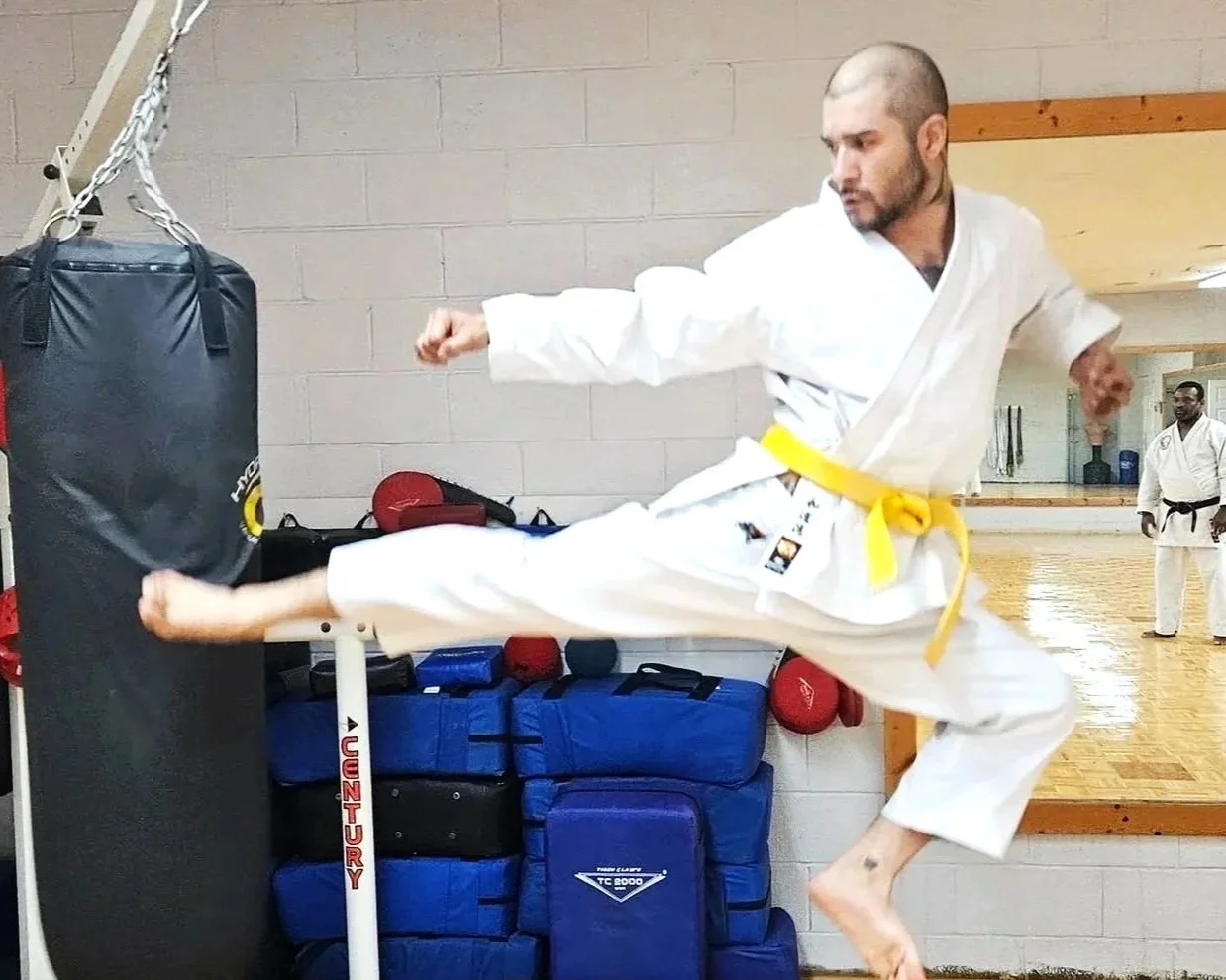 Martial artist practicing a flying kick in a dojo, wearing a white gi with a yellow belt, kicking a punching bag, with equipment and another person observing in the background.