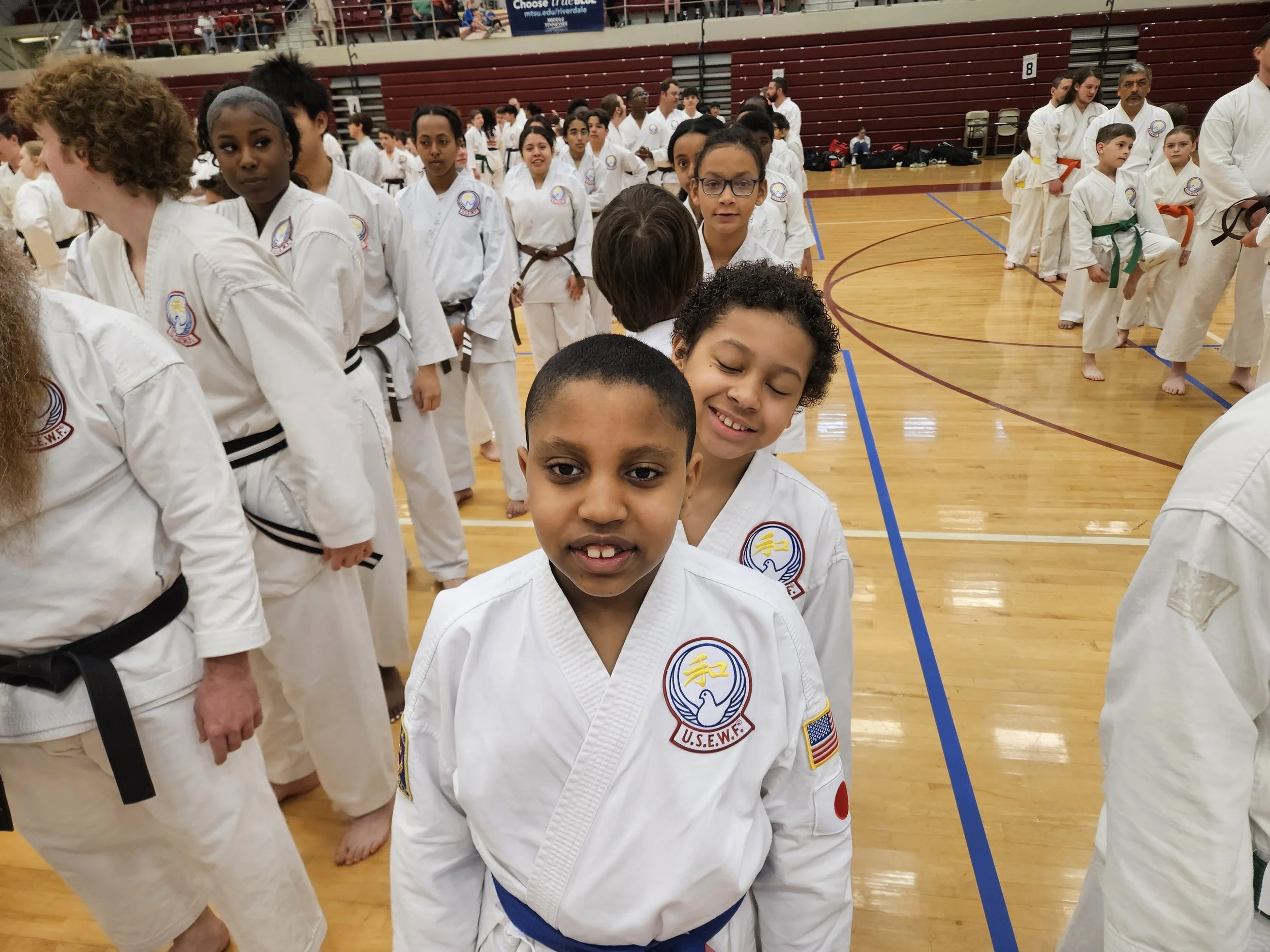 Children dressed in white martial arts uniforms standing in line on a gymnasium floor during a martial arts event.