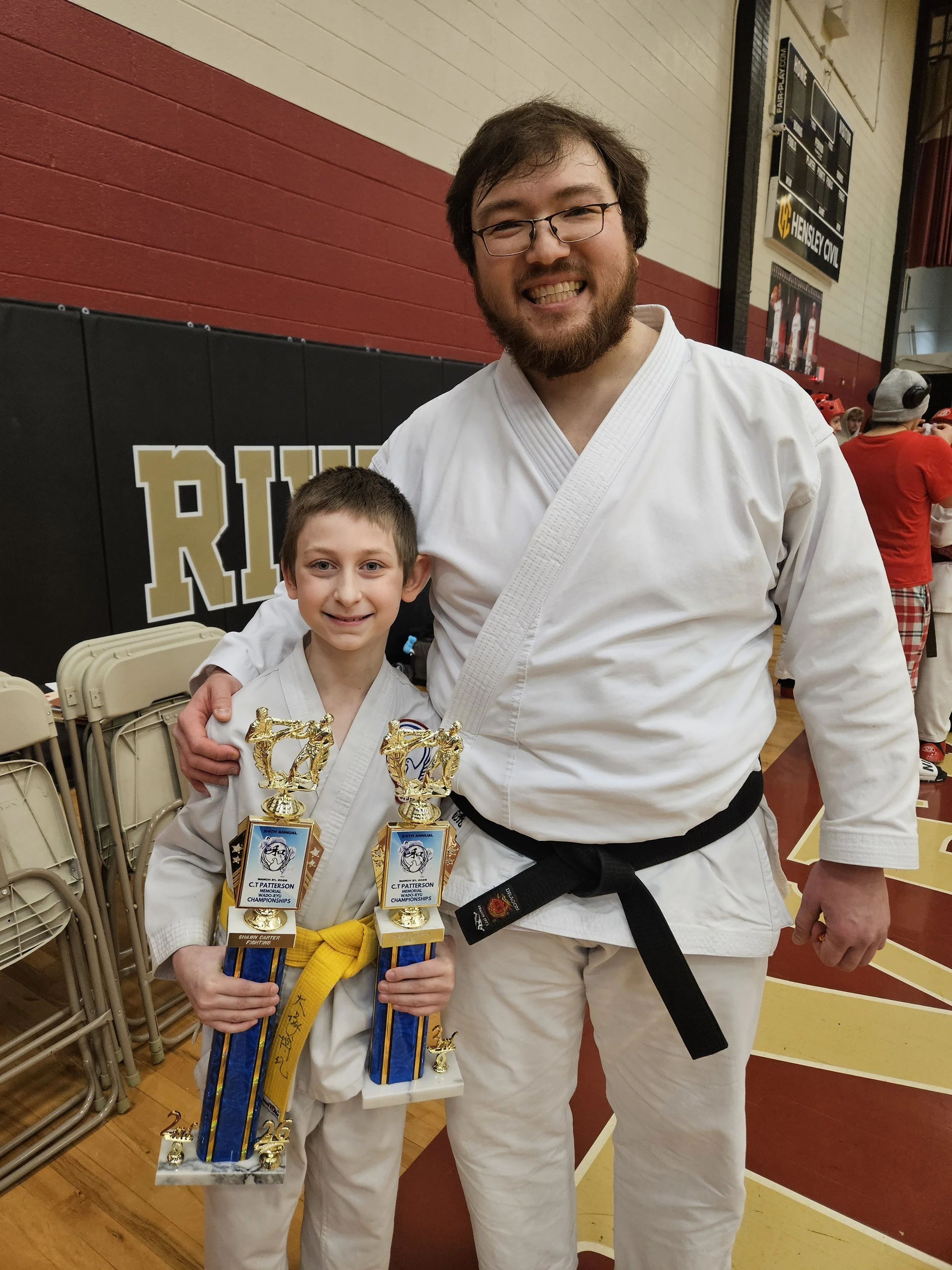 A young boy and an adult man in martial arts uniforms with black belts, smiling and holding trophies, in a gymnasium with red and beige walls and a scoreboard in the background.