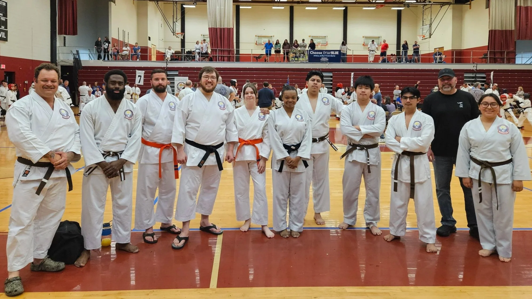 A group of martial artists in white gi uniforms with various colored belts standing in a gymnasium after a martial arts event, with spectators and other participants in the background.