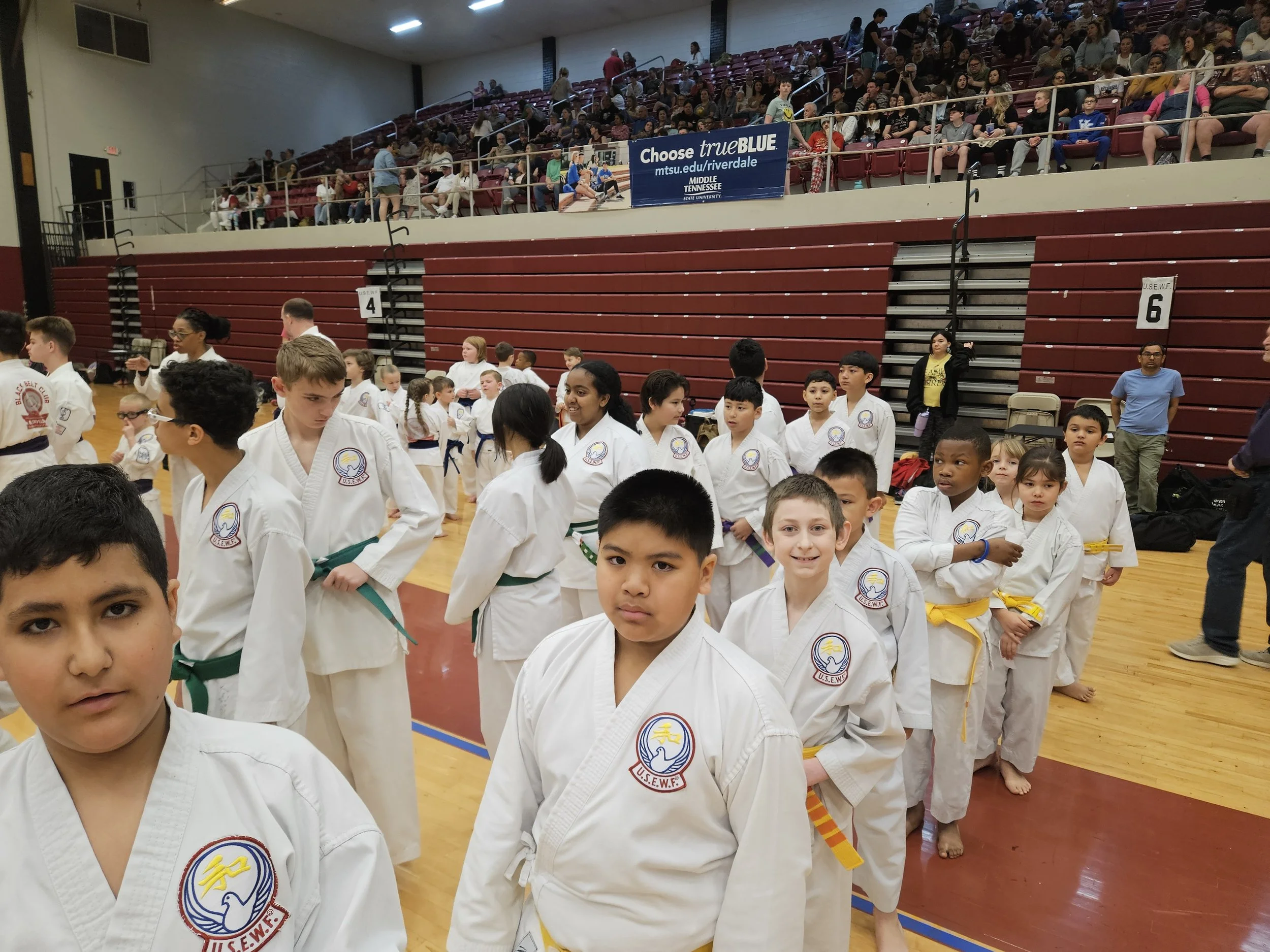 Young martial artists in white uniforms with colored belts in a gymnasium, many of them standing in line, some smiling, with spectators seated in the bleachers above.