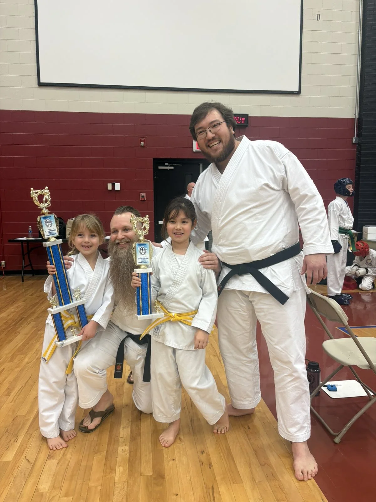 Group of four people in karate uniforms, two children and two adults, holding trophies, smiling after a karate competition in a gymnasium.