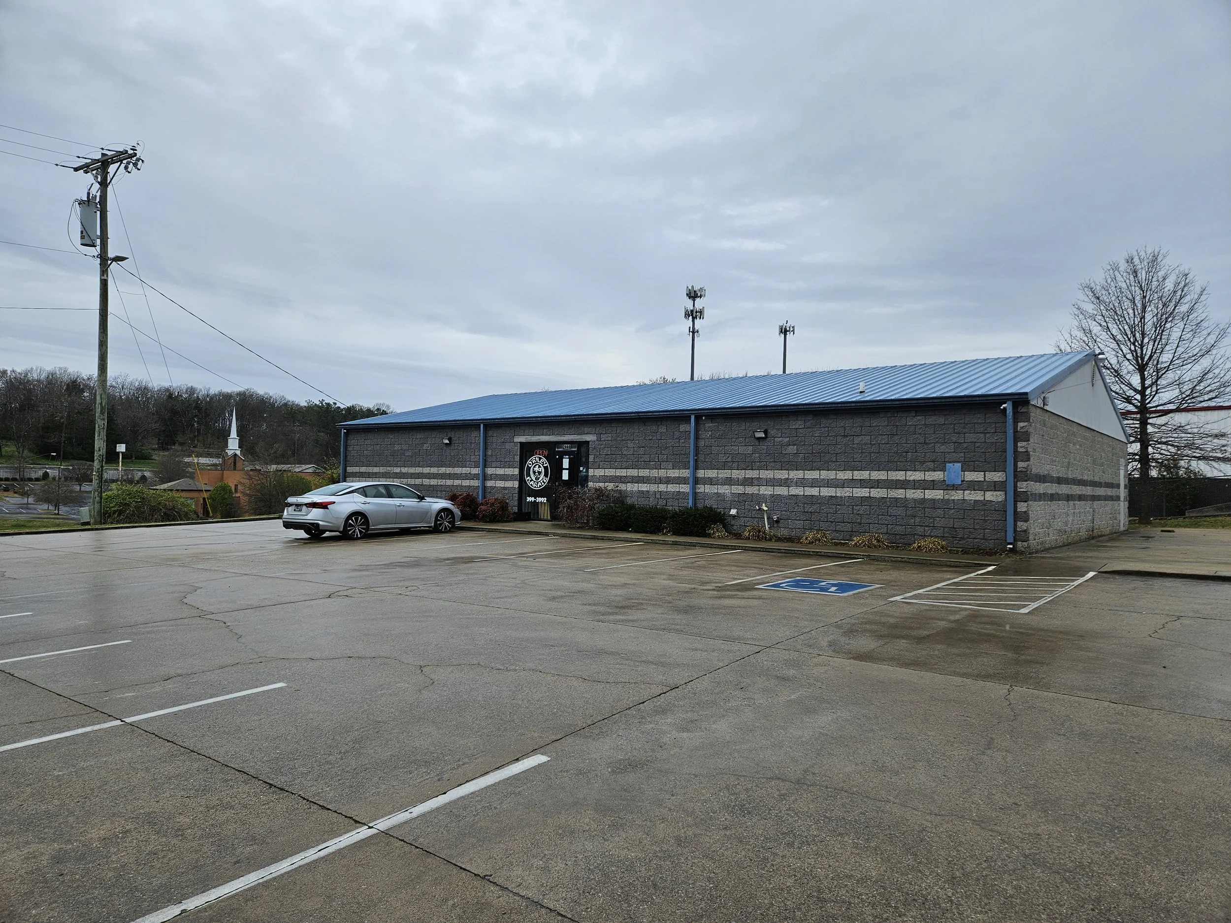 Empty parking lot with a single silver car near a gray brick building with a blue metal roof, and a designated handicapped parking space in the foreground, cloudy sky in background.