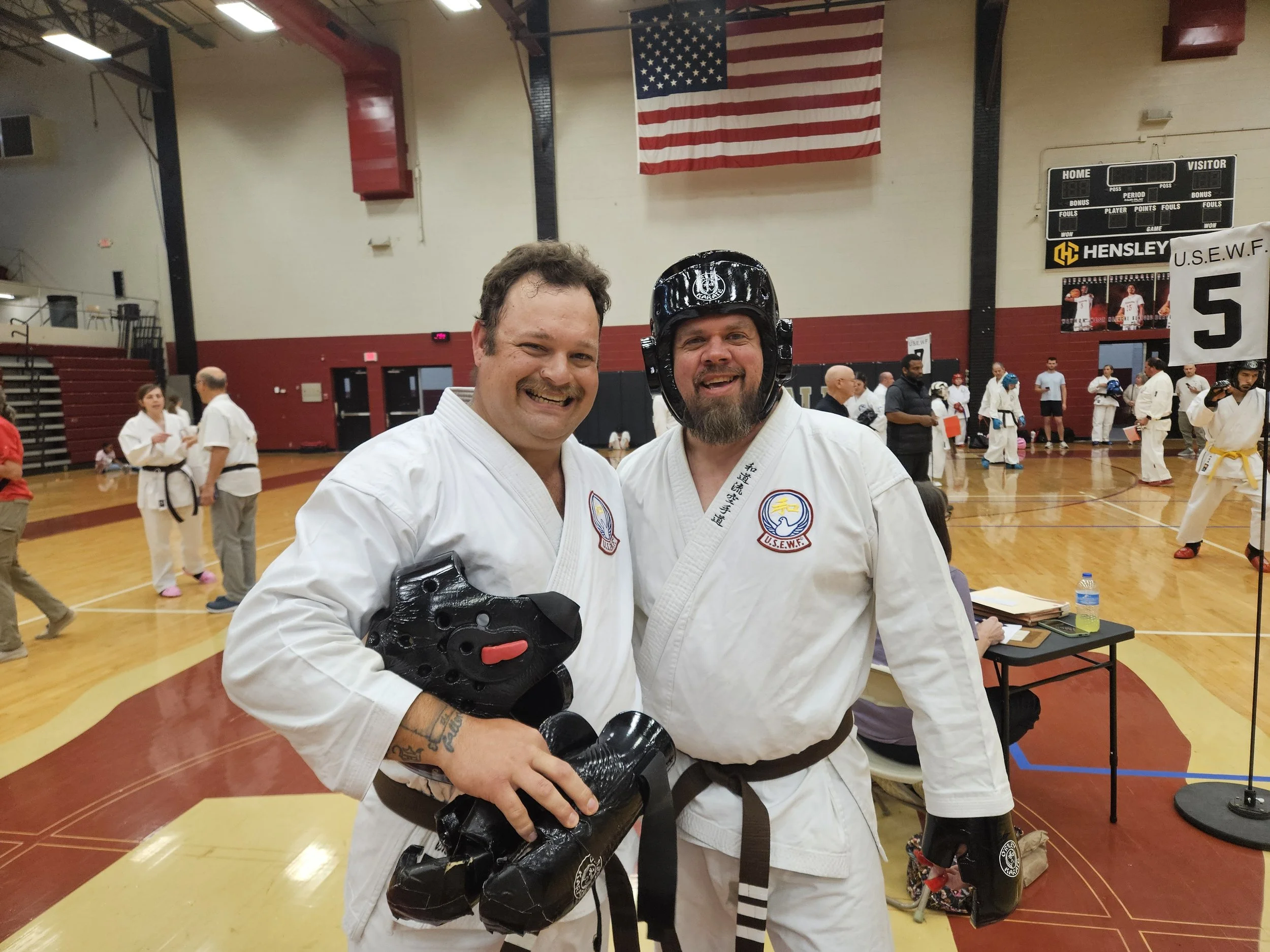 Two men in martial arts uniforms smiling at the camera in a gymnasium. One man holds black protective gear. Background shows other martial artists, some standing and some practicing. An American flag hangs on the wall.