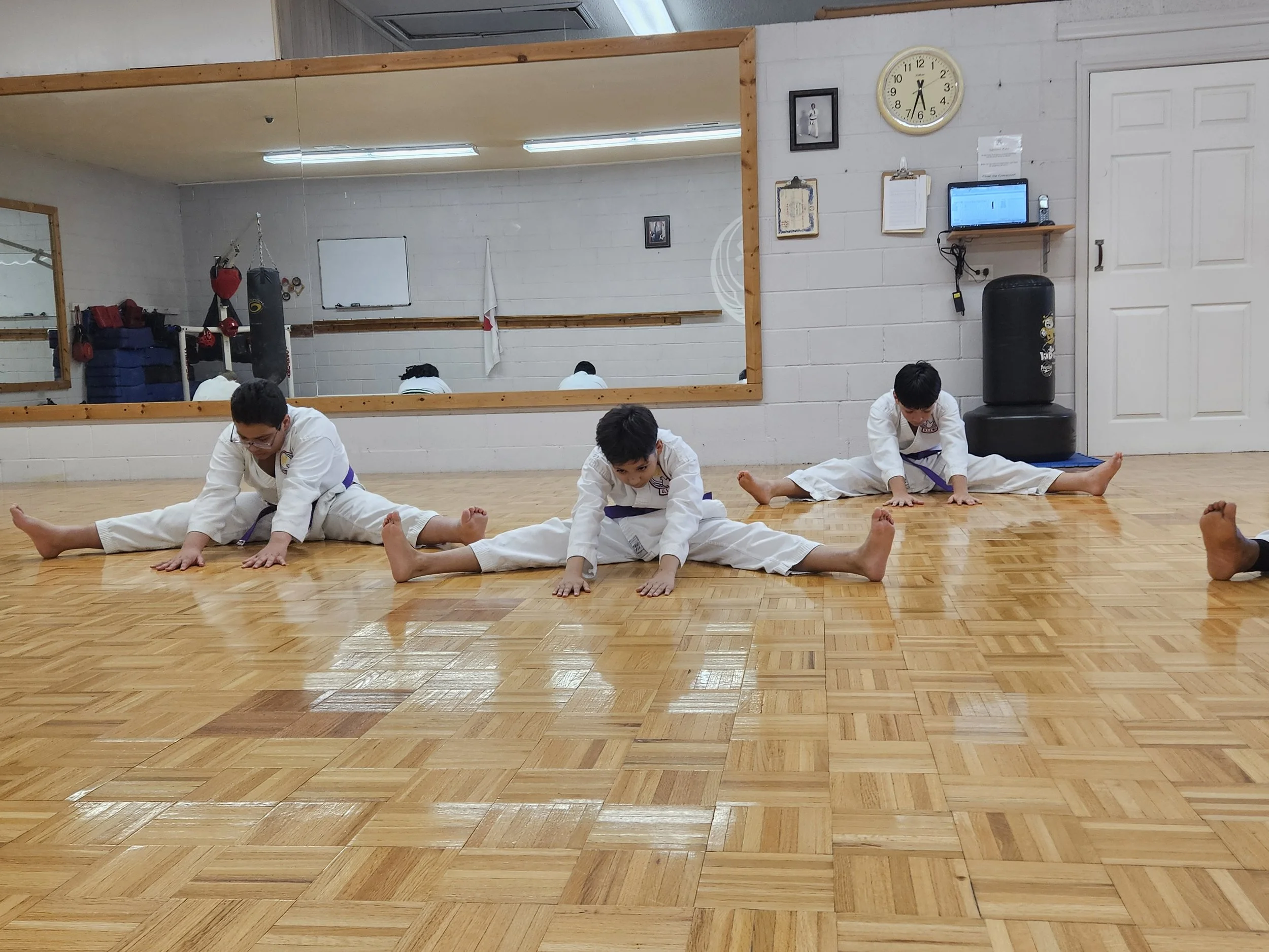 Three children in martial arts uniforms practicing stretching exercises on a hardwood floor in a dojo or gym, with a large mirror, clock, and equipment in the background.
