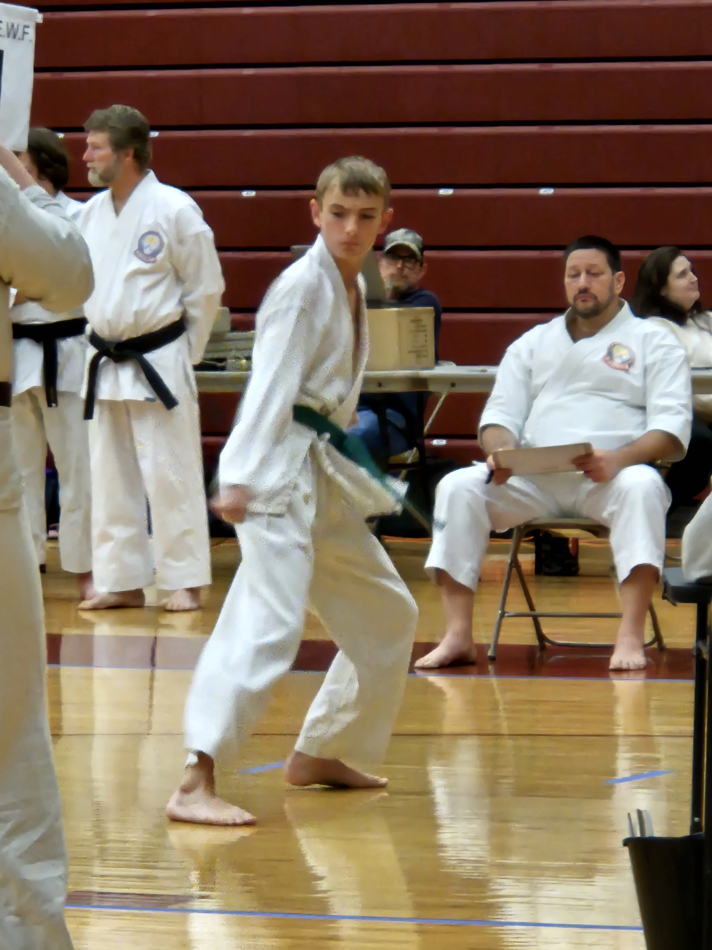 Young martial artist in a gi practicing a move during a martial arts event, with officials and spectators seated behind.