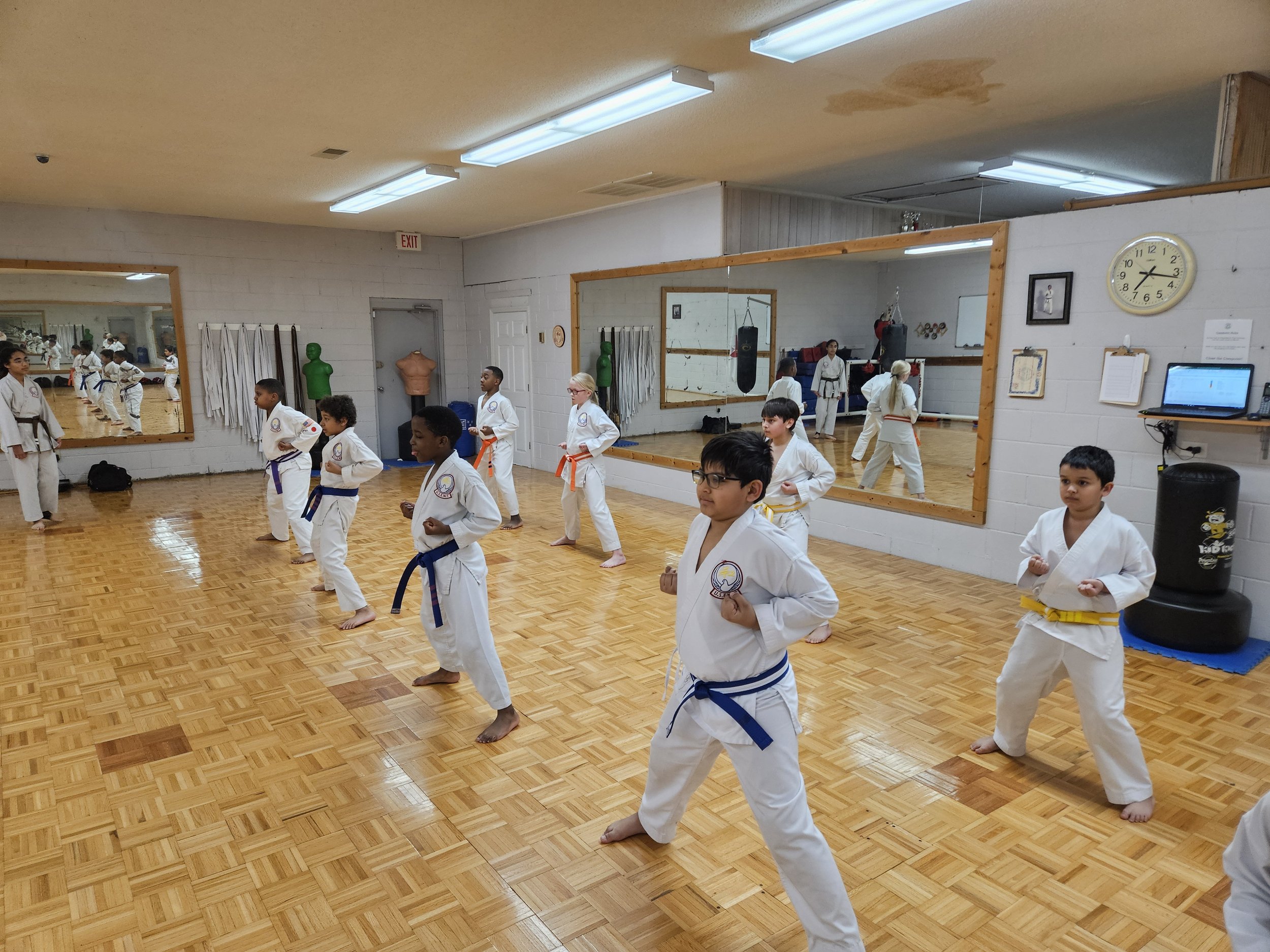 Children practicing taekwondo in a martial arts class, wearing white uniforms and colored belts, in a room with hardwood floors, large mirrors, and punching bags.
