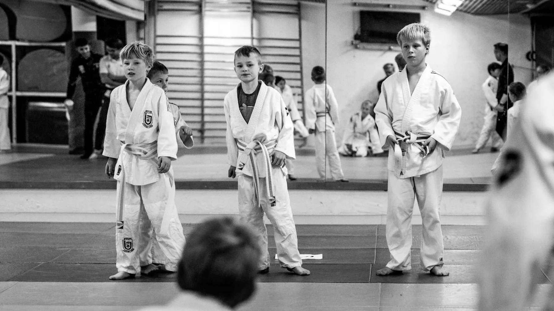 Children in judo uniforms standing on a mat during a judo class or competition, with a mirror and other children practicing in the background.