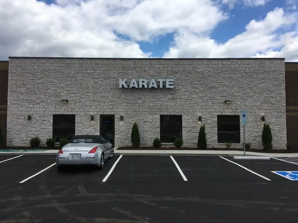A karate school with a brick facade, three windows, and a sign that reads 'KARATE' on the front. There is a parking lot with a silver car parked in front and a handicap parking space nearby.