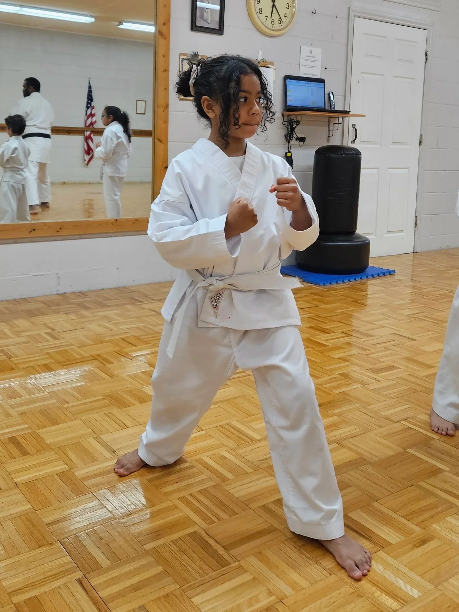 A young girl practicing karate in a dojo, wearing a white karate gi with a belt, standing in a ready stance with fists clenched, in front of a mirror and a punching bag, with others training in the background.